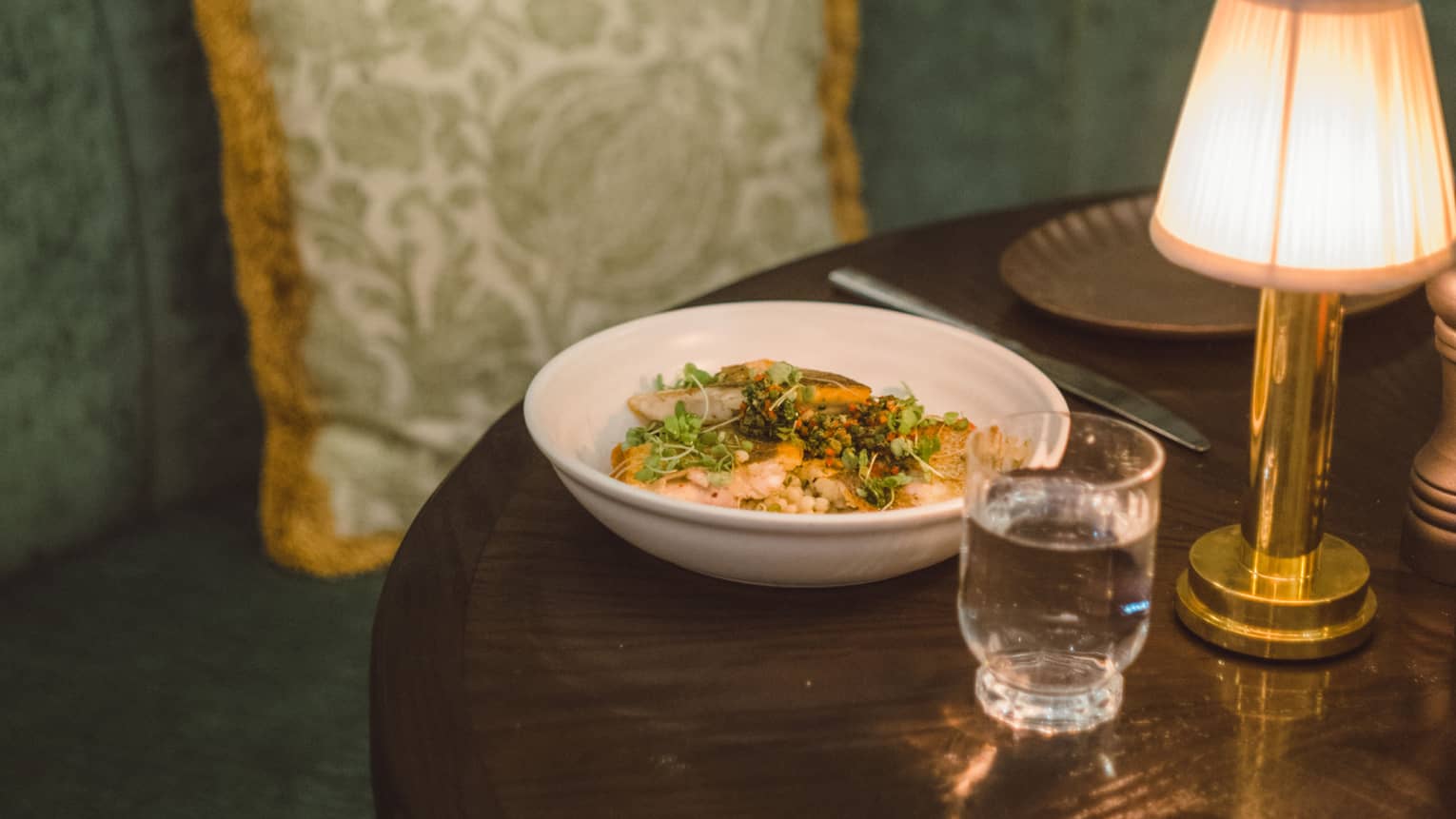 A colourful dish sits atop a wooden, lamplit table with a green velvet booth behind