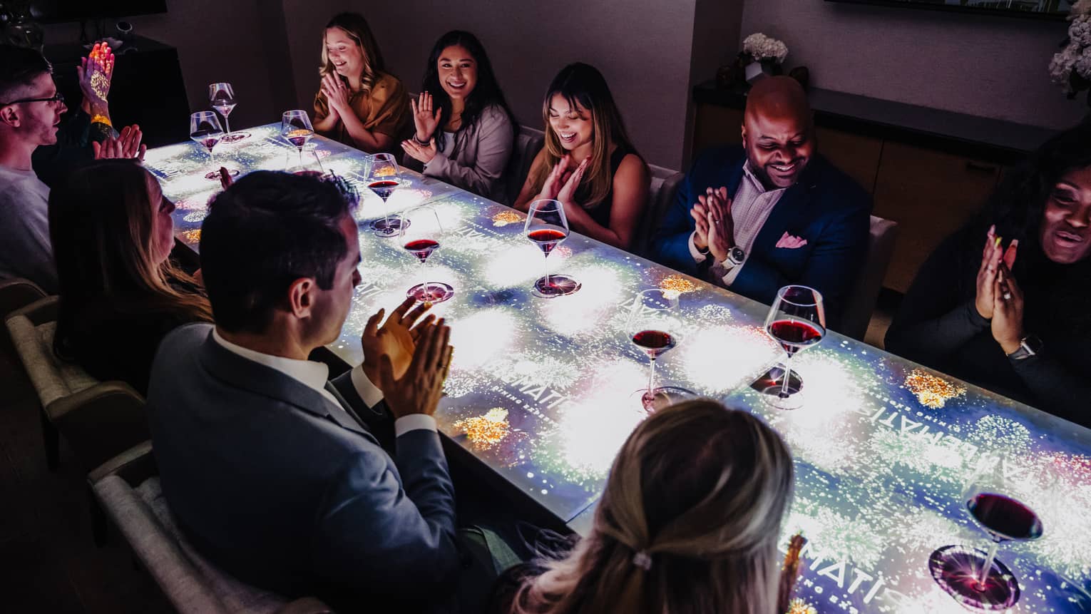 A large group of people sitting at a table with a screen on top of it drinking wine.