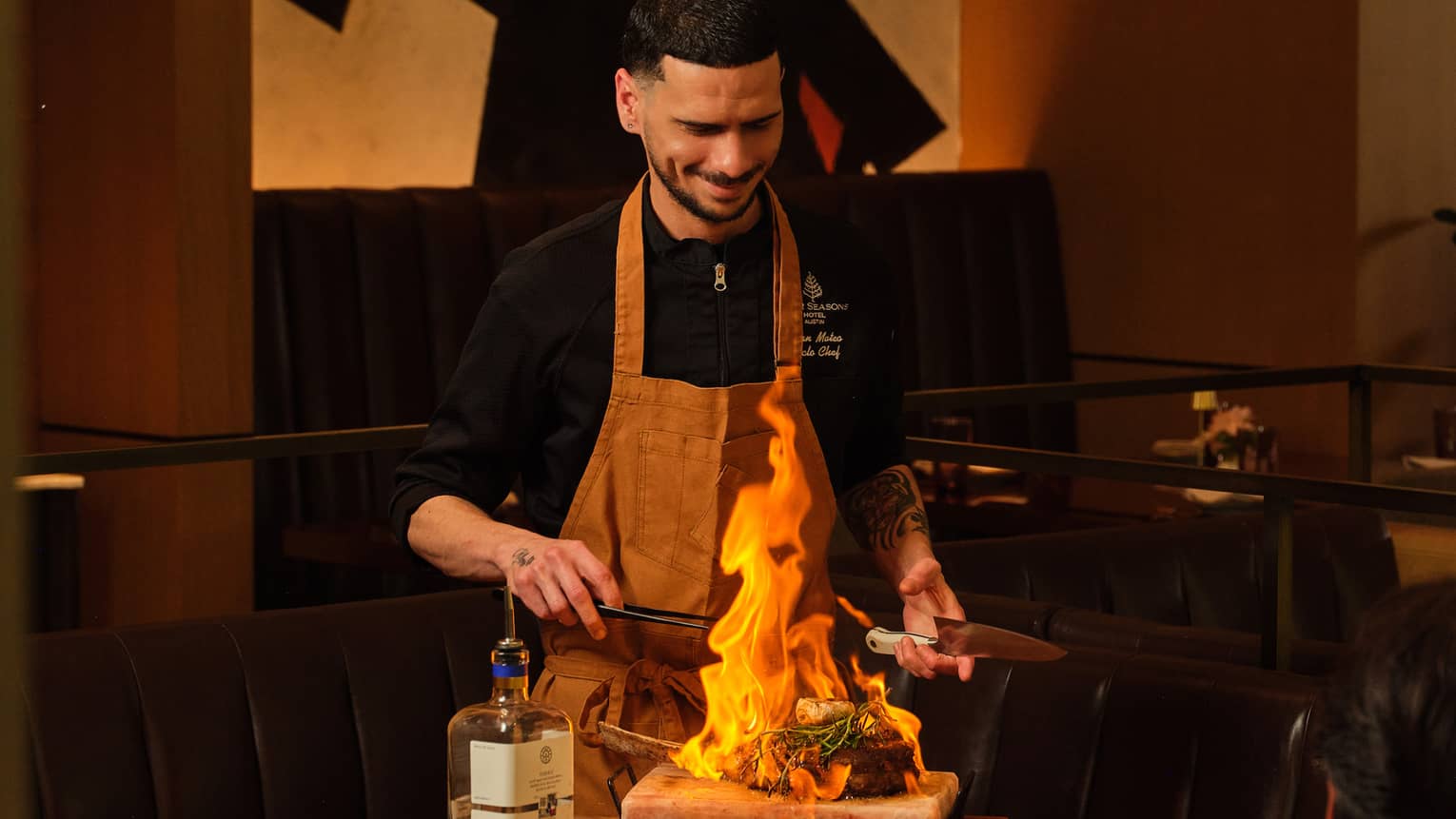A chef stands in front of a flaming dish on a butcher block?style food prep table in an indoor dining space