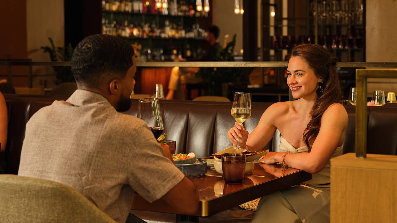 Two people drink beverages from stemmed glasses, sitting across from each other at a dining table in a serene and cozy indoor dining space.