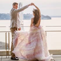 A groom in a tan suit twirls a bride dressed in a light pink dress on a terrace overlooking the pier and the Seattle Great Wheel.
