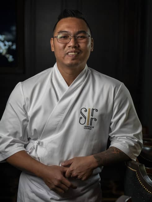 Chef with short dark hair and glasses, wearing a white chef's jacket and apron, stand smiling at the camera