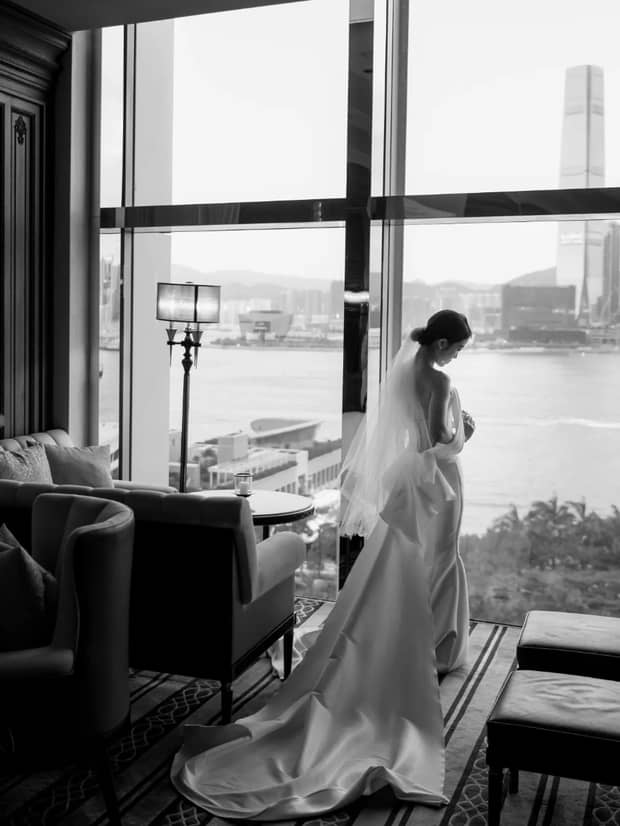 A woman dressed in a silk wedding dress with a long train stands in a sitting room by a large window overlooking the city of Hong Kong