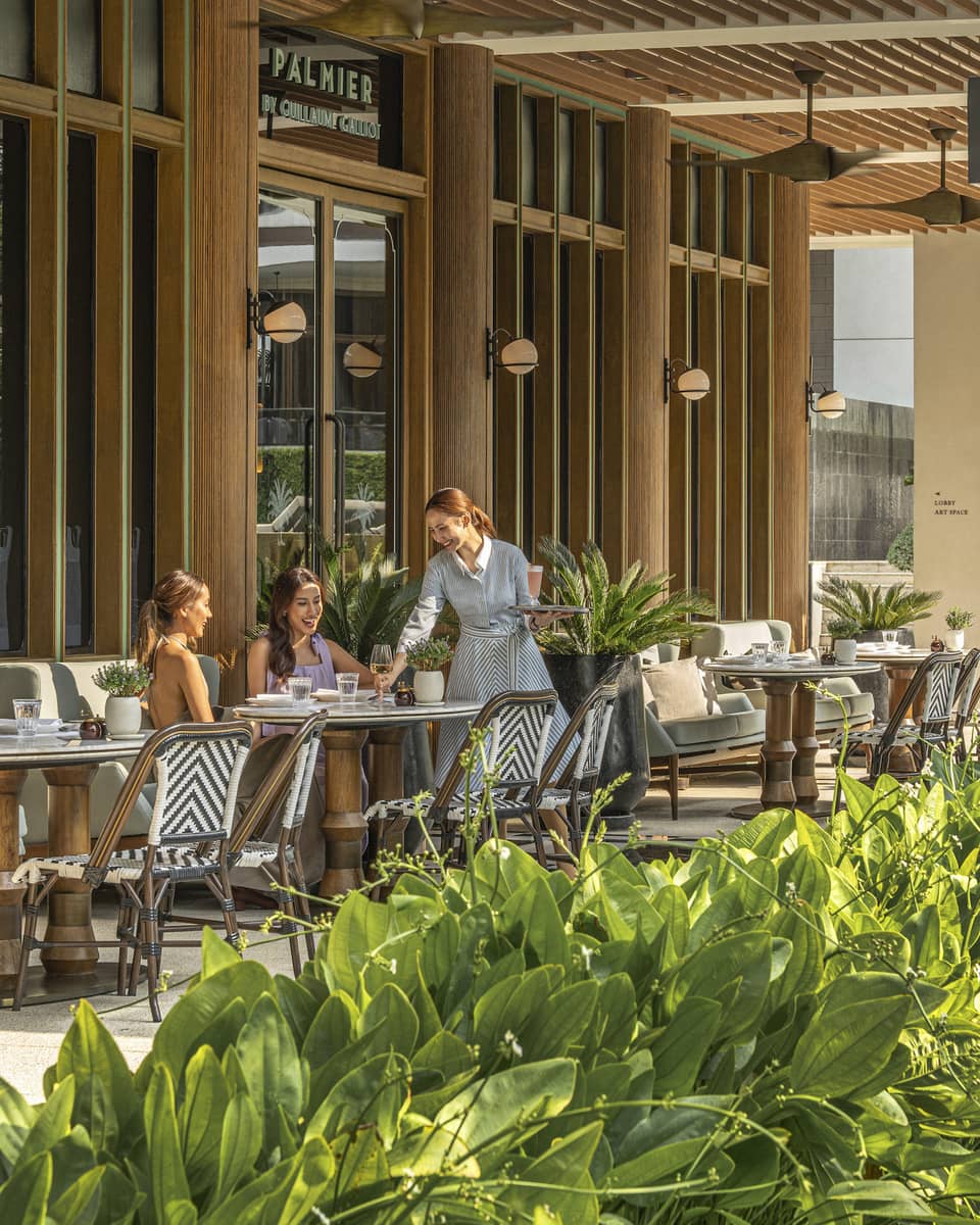 Two dining patrons are served by a waiter on the shaded patio of a restaurant