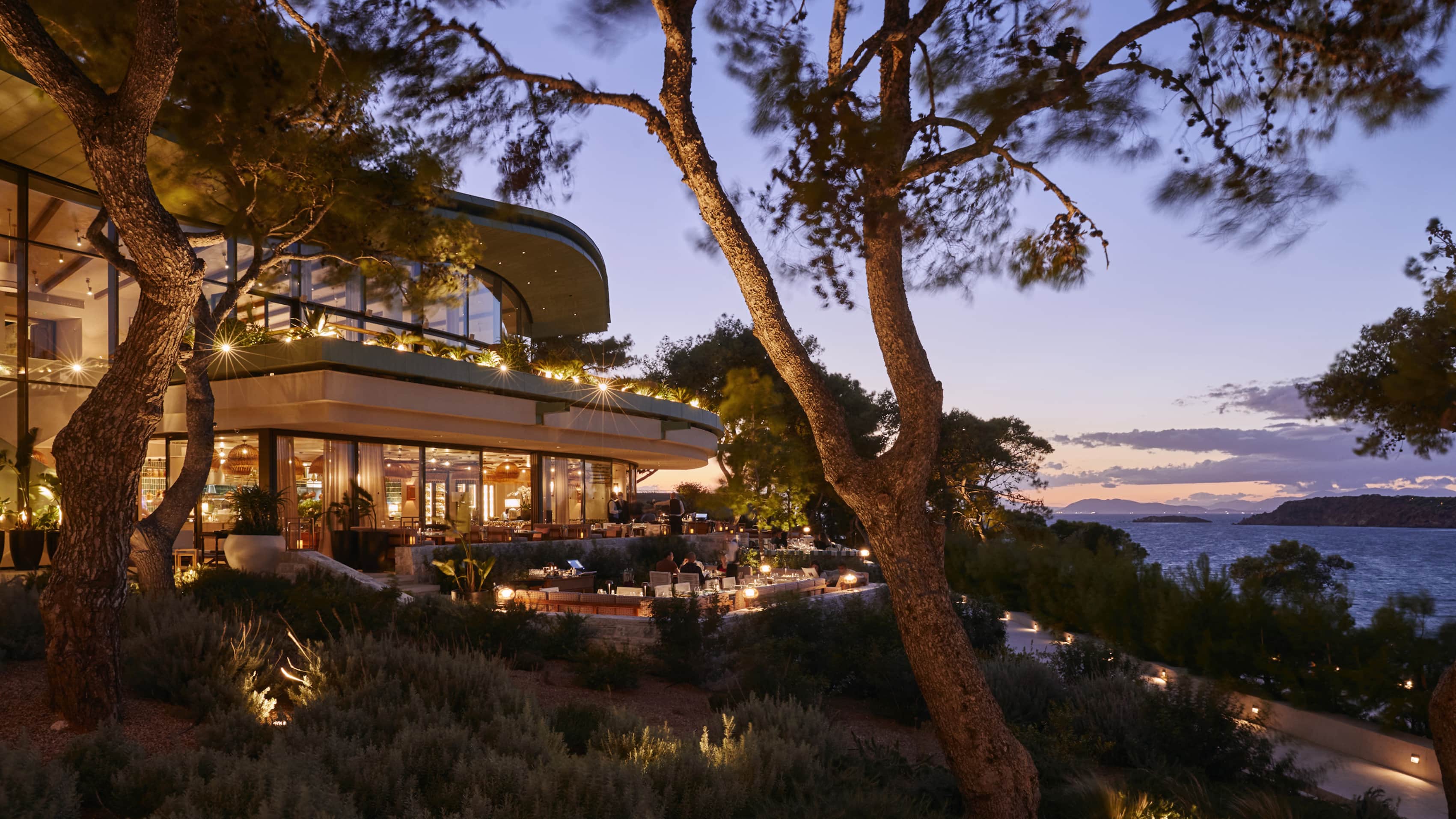 Beefbar on the Astir peninsula at dusk, a lit glass pavilion under Aleppo pines above the water.