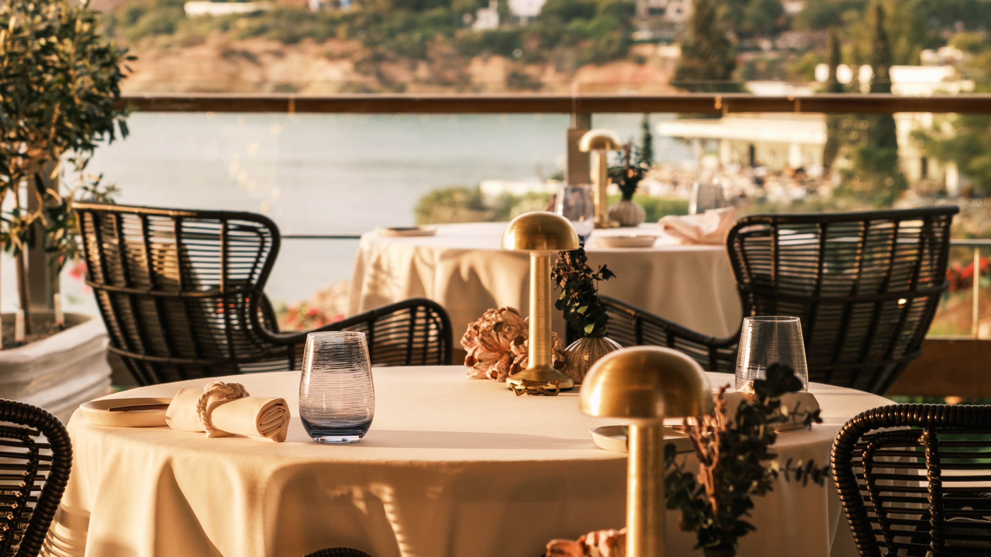 The terrace at Pelagos: round tables in cream linen, black rattan chairs and brass lamps, the bay behind.