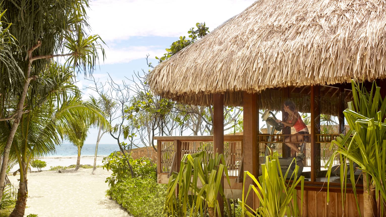 Woman on exercise bike in thatched-roof gym looks out at beach