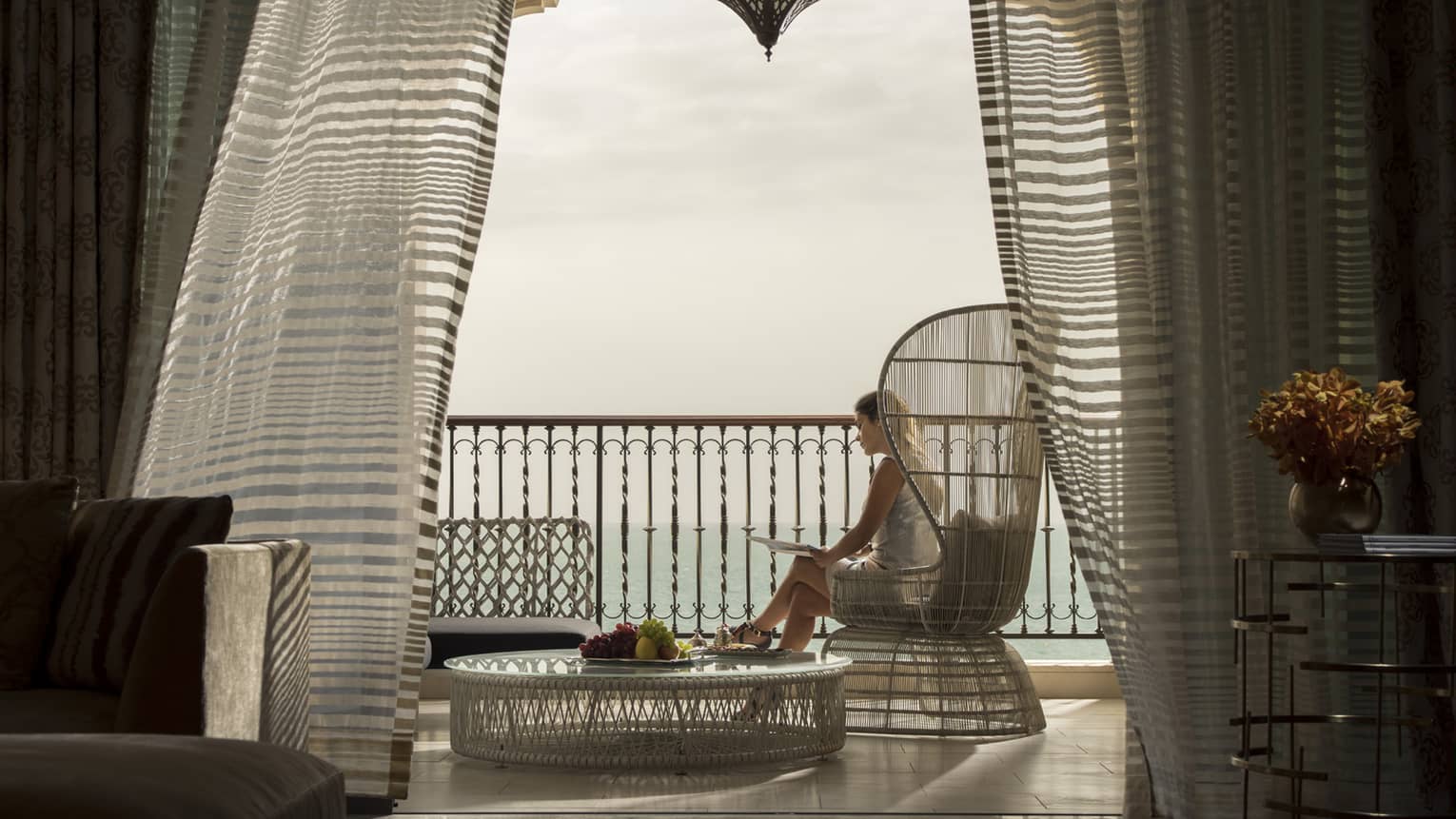 Woman sits, reads in tall wicker chair on patio beside wicker coffee table with fresh fruit