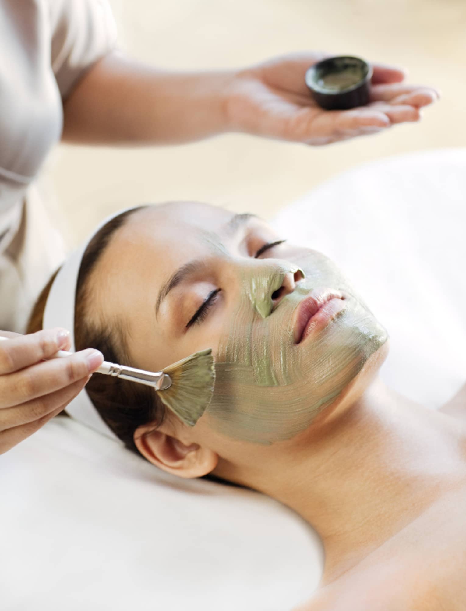 Spa staff holding brush paints green mask on woman's cheeks, chin, nose 