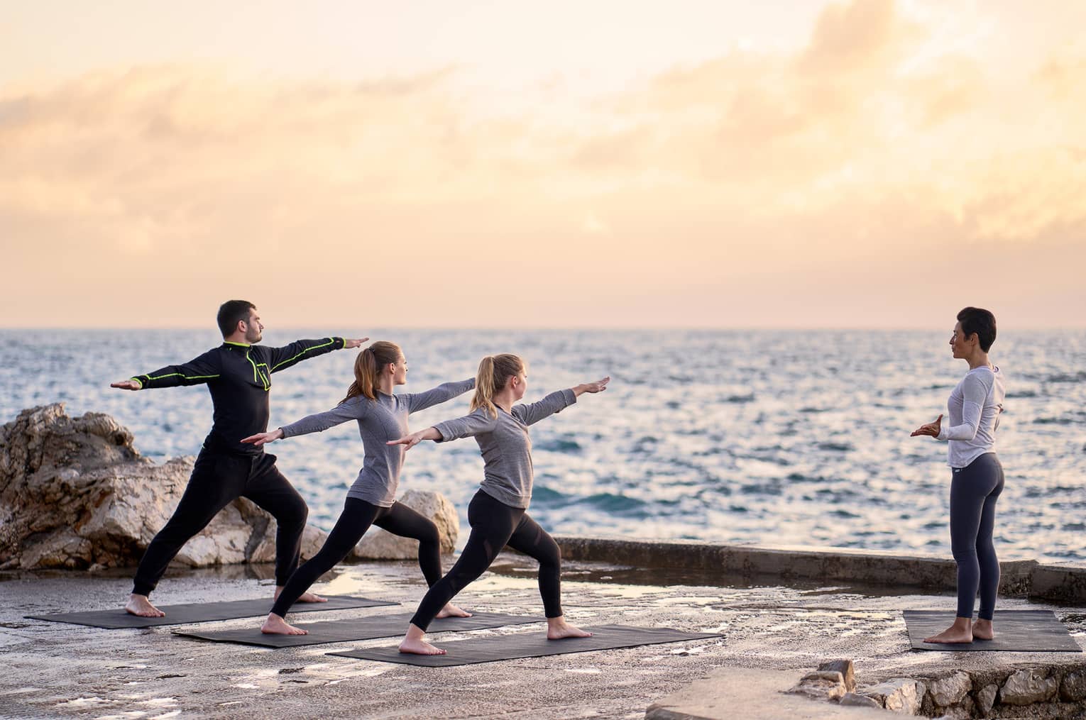 Three people in warrior yoga pose and coach standing at sea's edge at sunrise