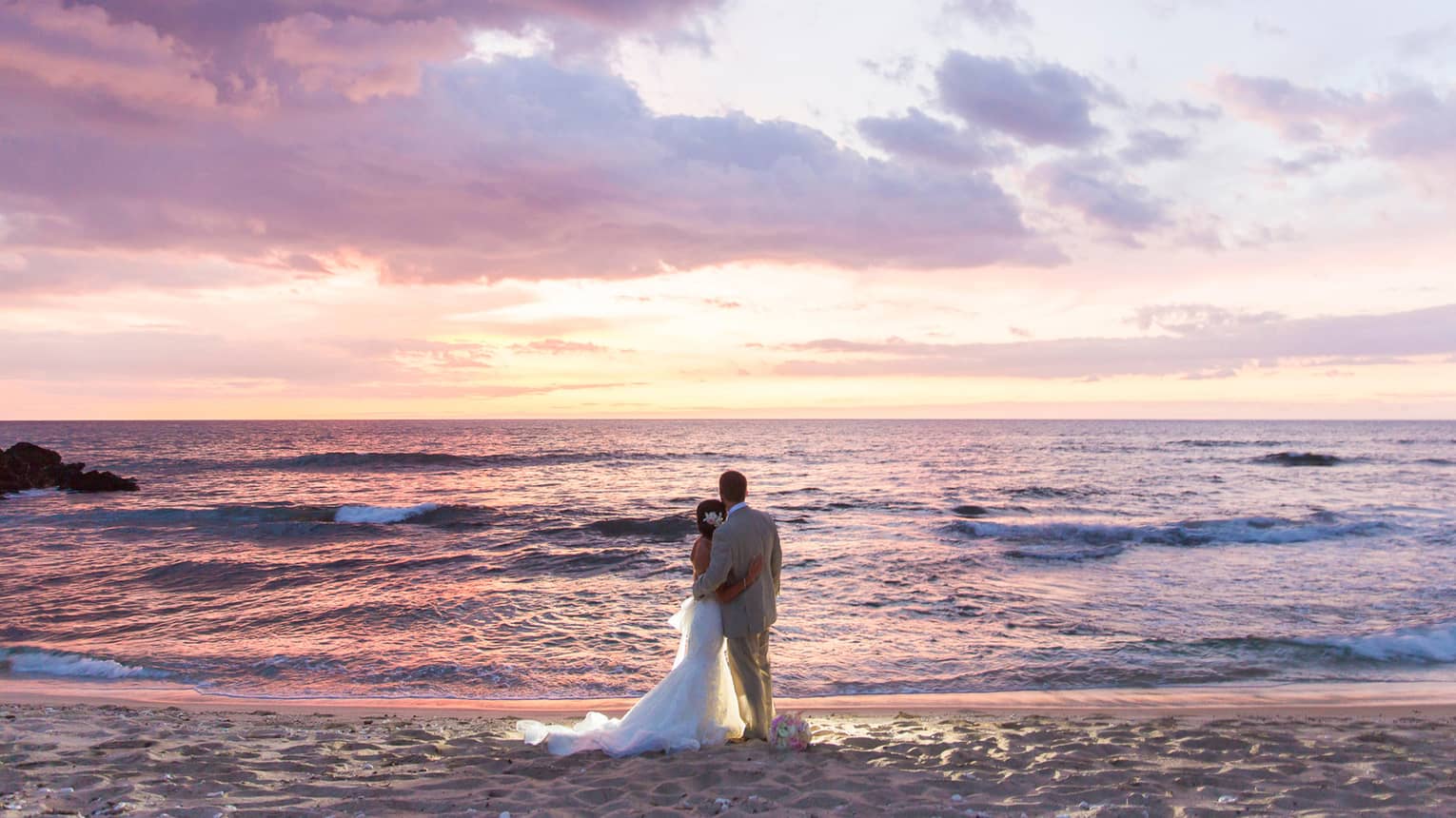 Bride and groom stand on sand beach and watch sunset over ocean