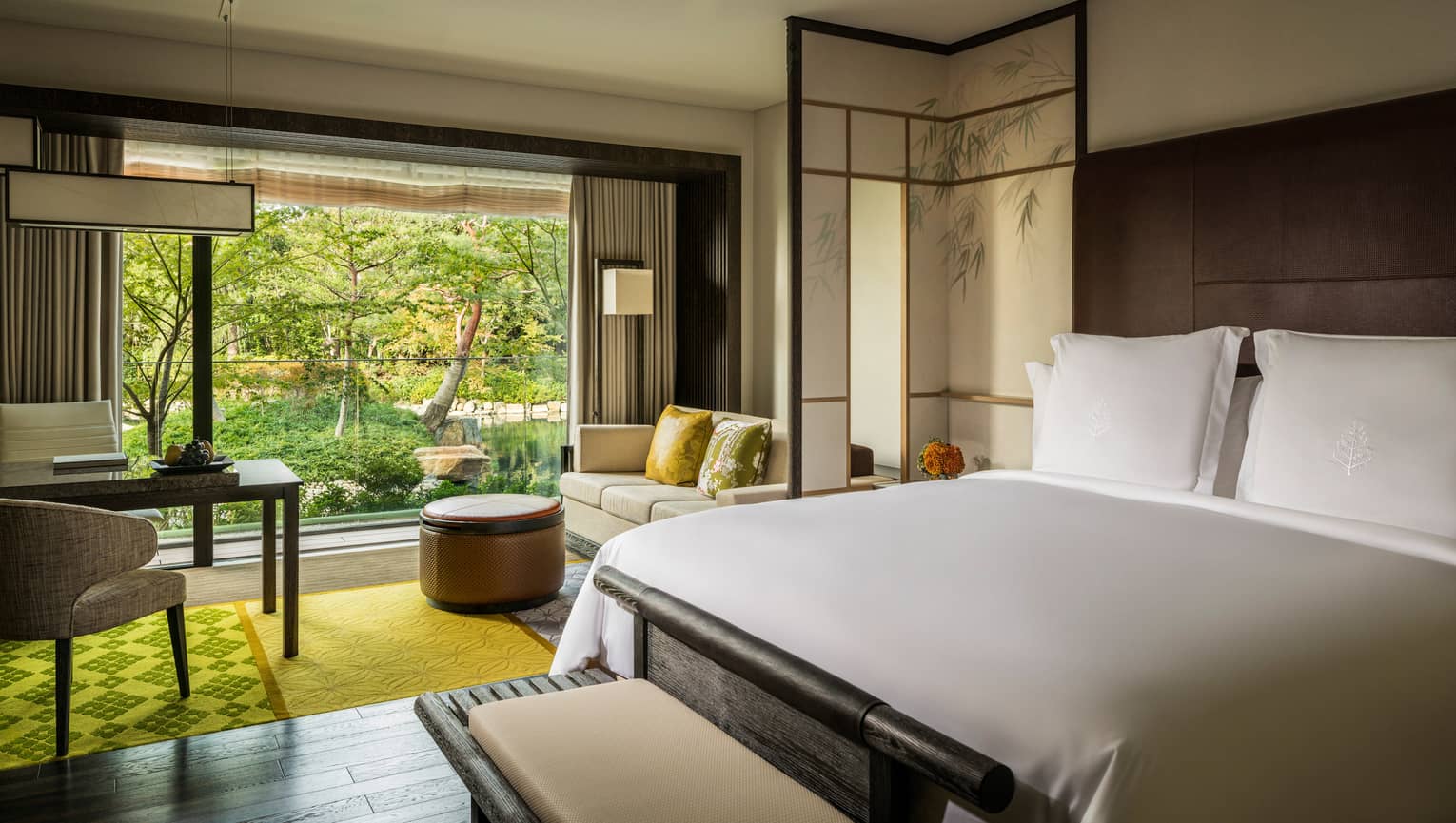 Guest room with a king bed, sofa with green cushions, round ottoman and large window overlooking a pond garden