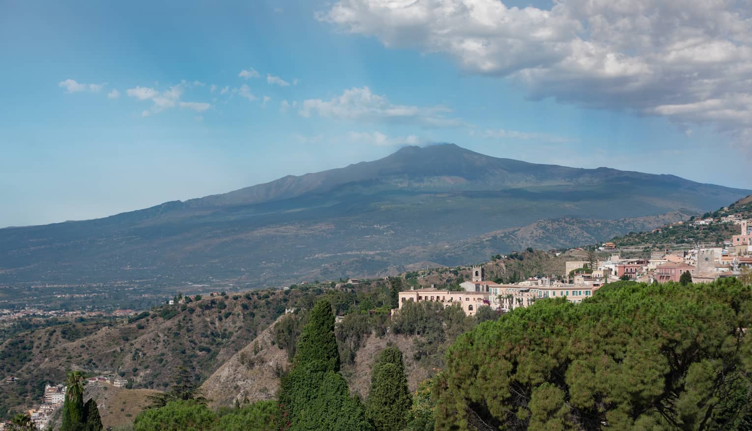 Hillside buildings surrounded by trees, Mount Etna and blue sky in backdrop