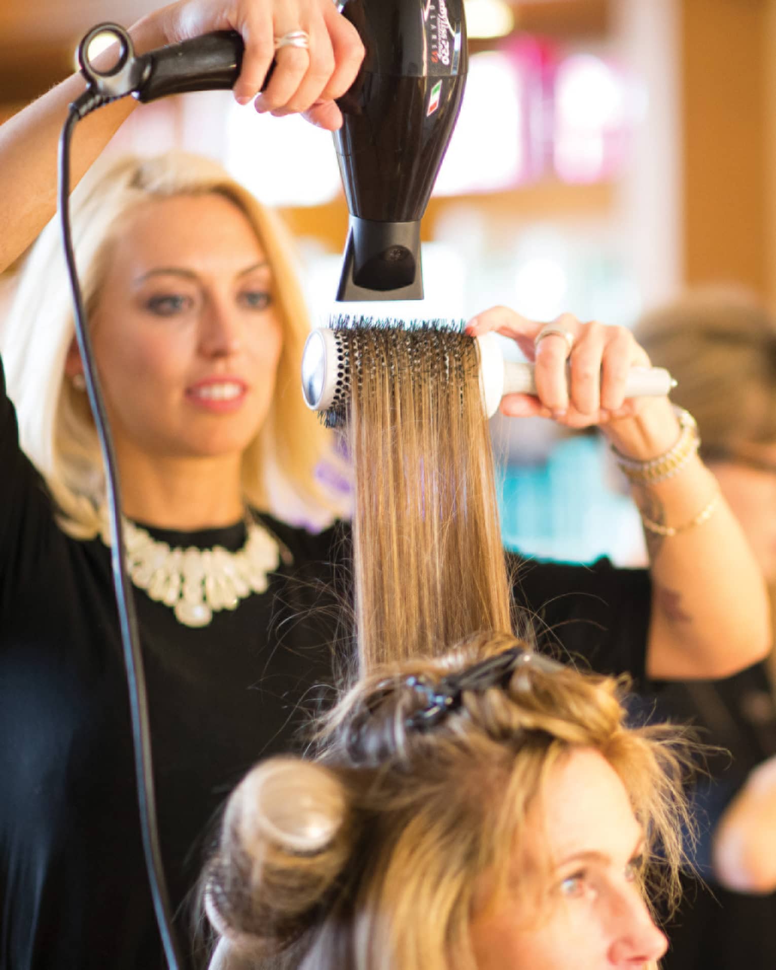 Hairstylist with hairdryer pulls back woman's hair with round brush in salon