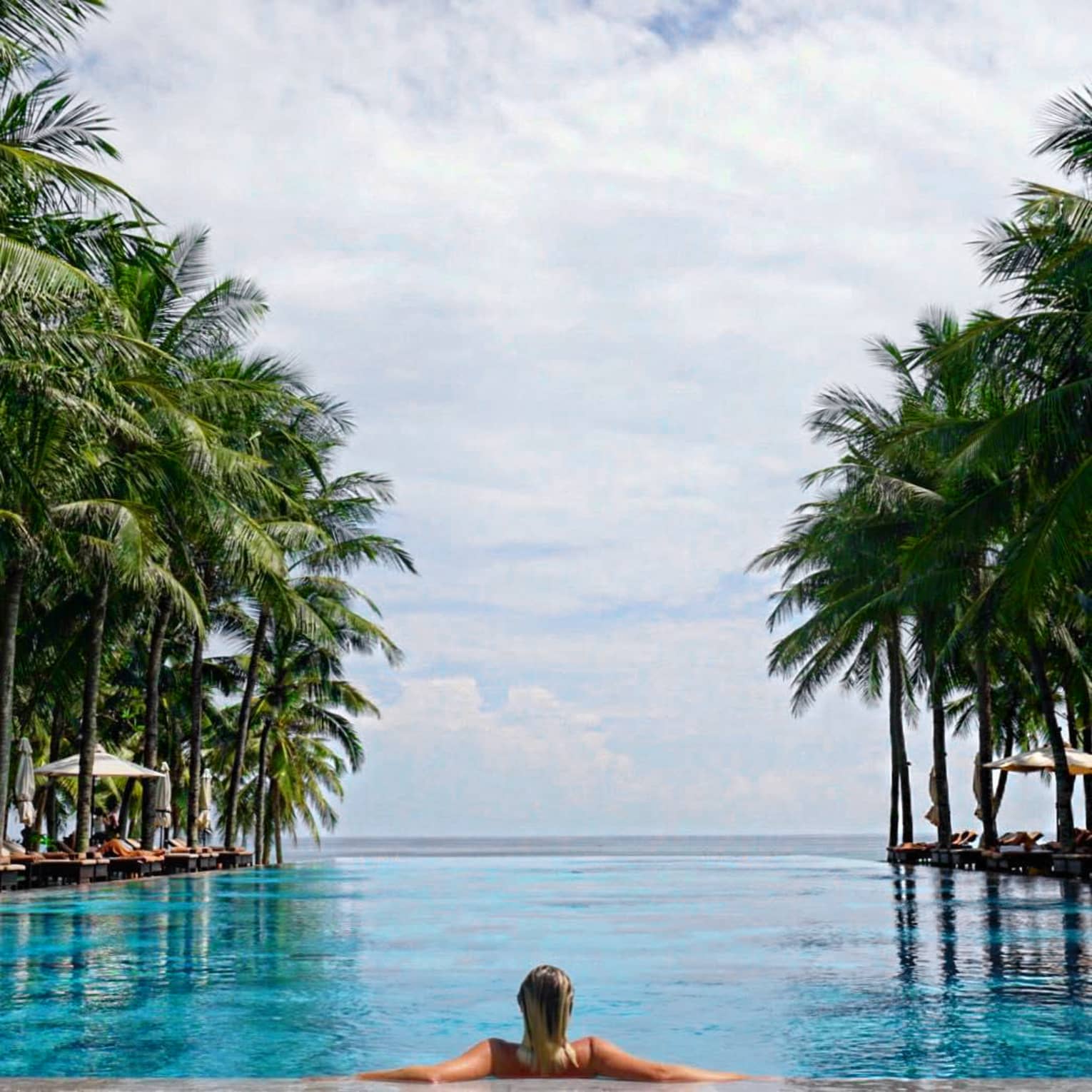 Back of woman leaning against ledge of long outdoor swimming pool under large palms