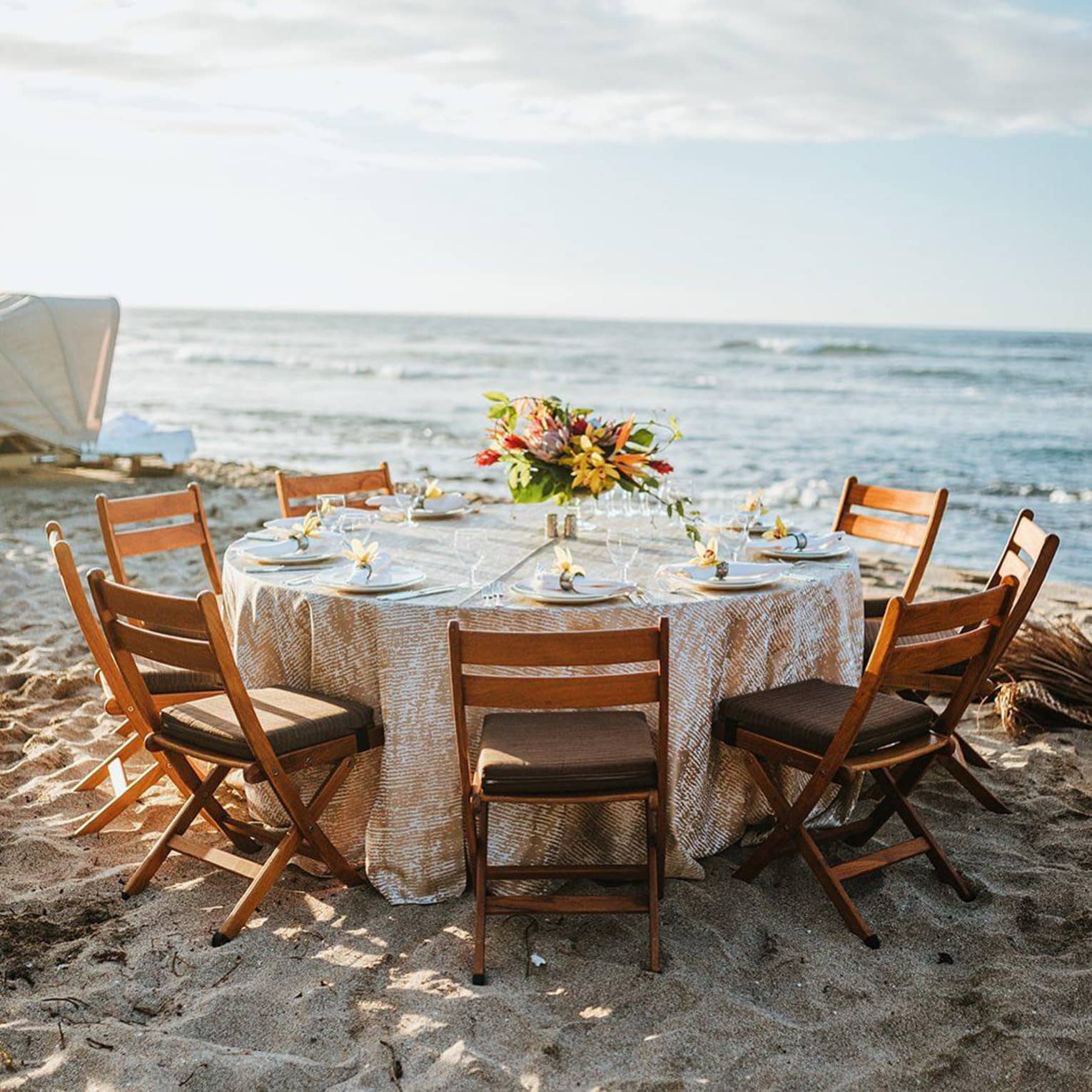 A wedding reception table adorned with glassware and a floral centerpiece is set up on a sandy beach by the ocean. 