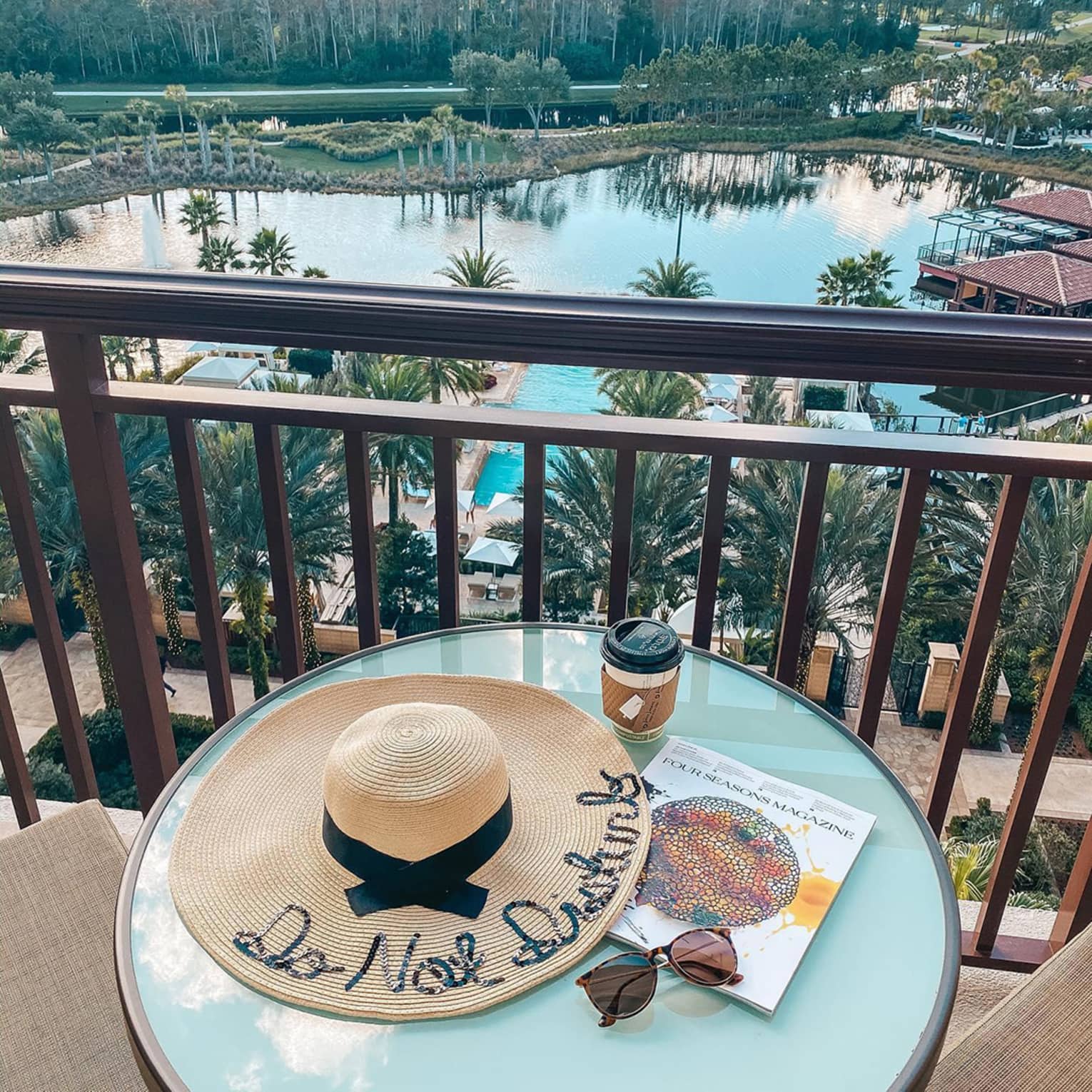 Beach hat with words Do Not Disturb, magazine, glasses and coffee on balcony table overlooking swimming pool