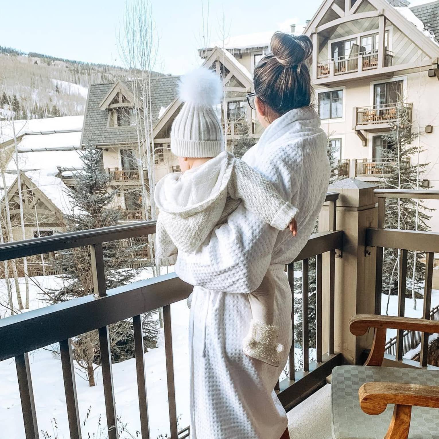 ,A woman dressed in a white robe and slippers holds a small child on a balcony overlooking a snow capped hotel