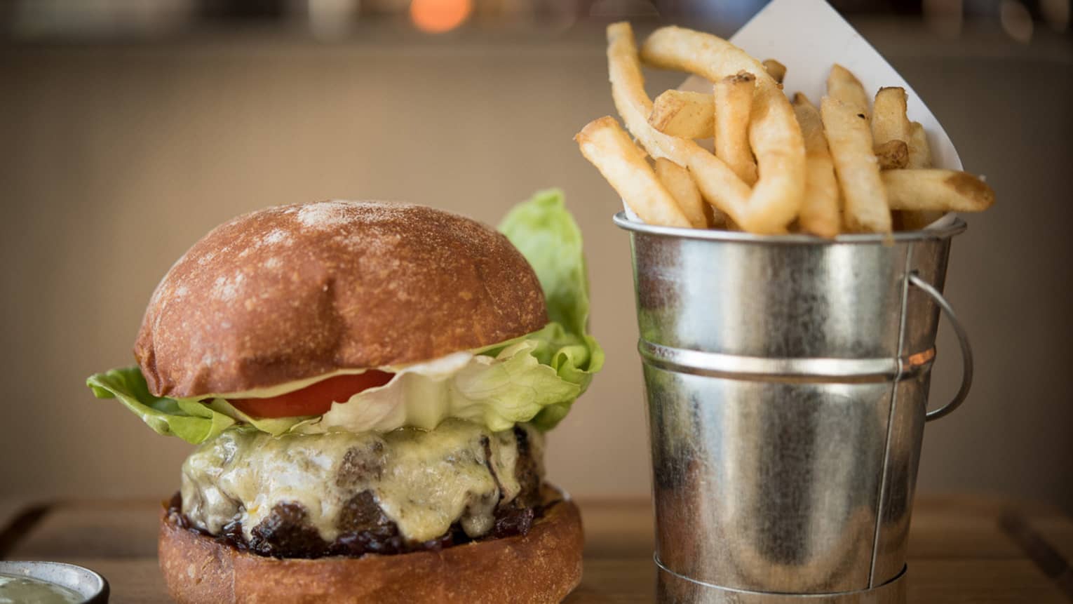 Thick beef burger with melted cheese on artisan bun beside silver tin with French fries