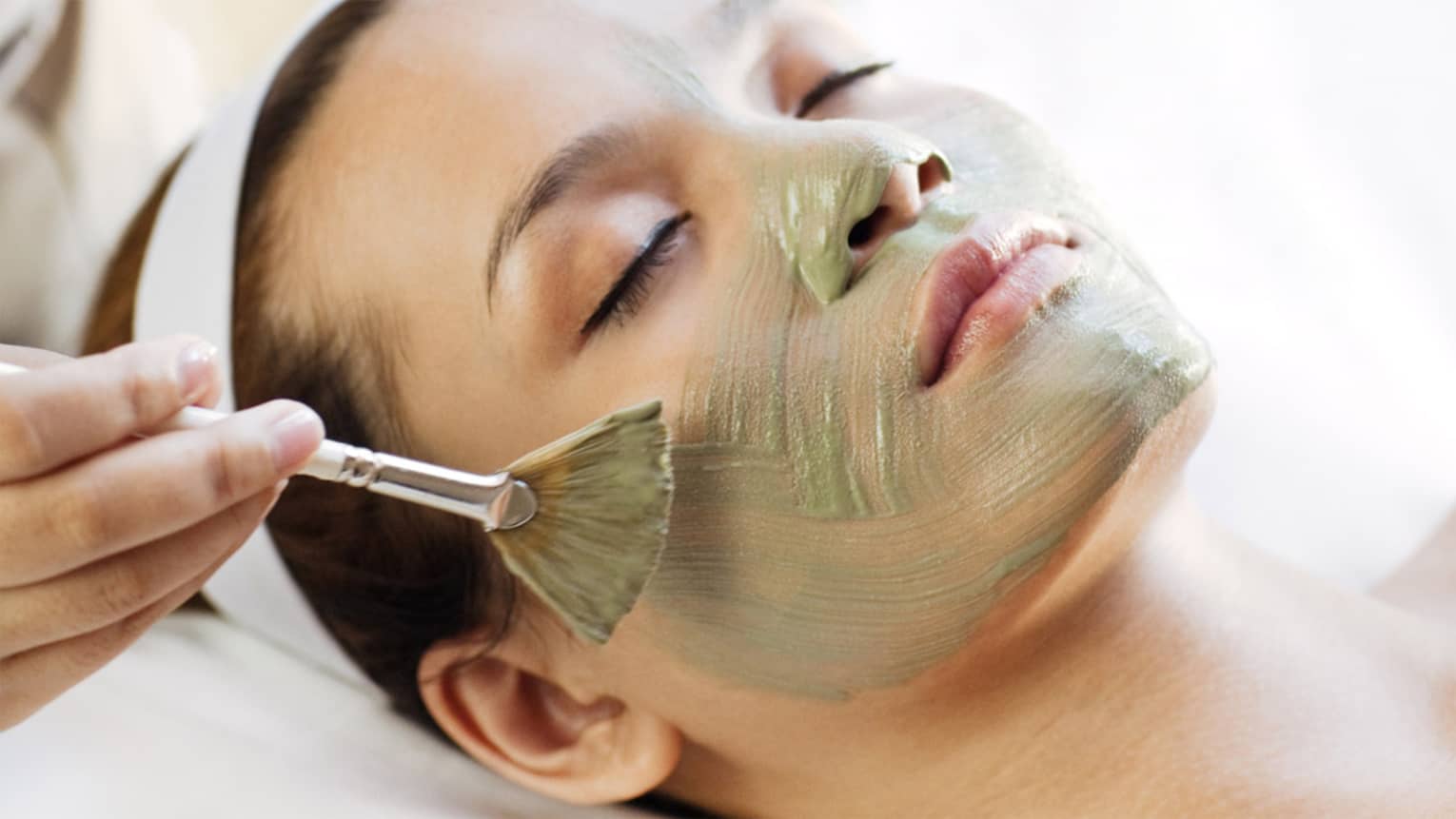 Spa staff holding brush paints green mask on woman's cheeks, chin, nose 