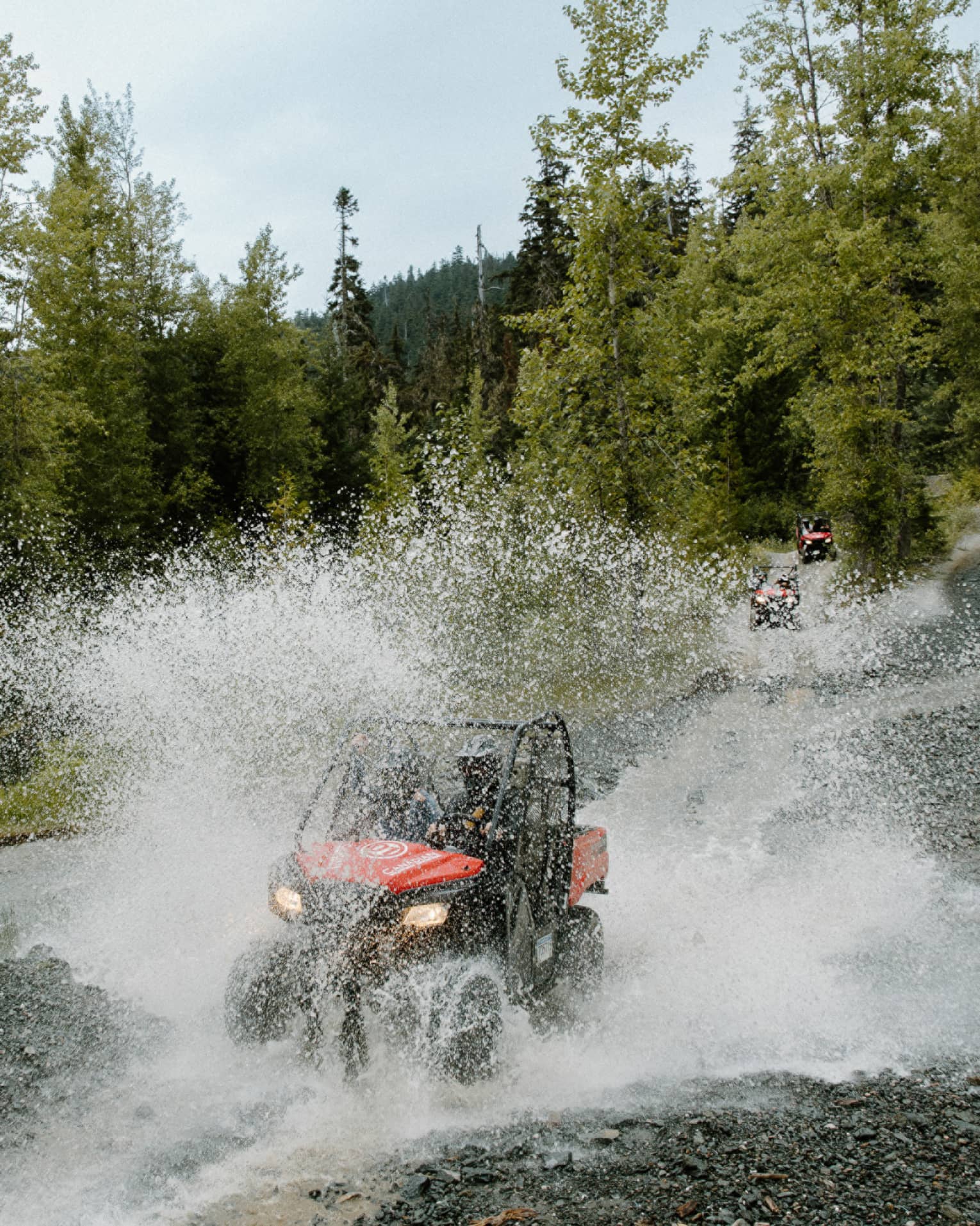 A red-and-black ATV driving through water, creating a thick veil of water spray against a background of pine forest.