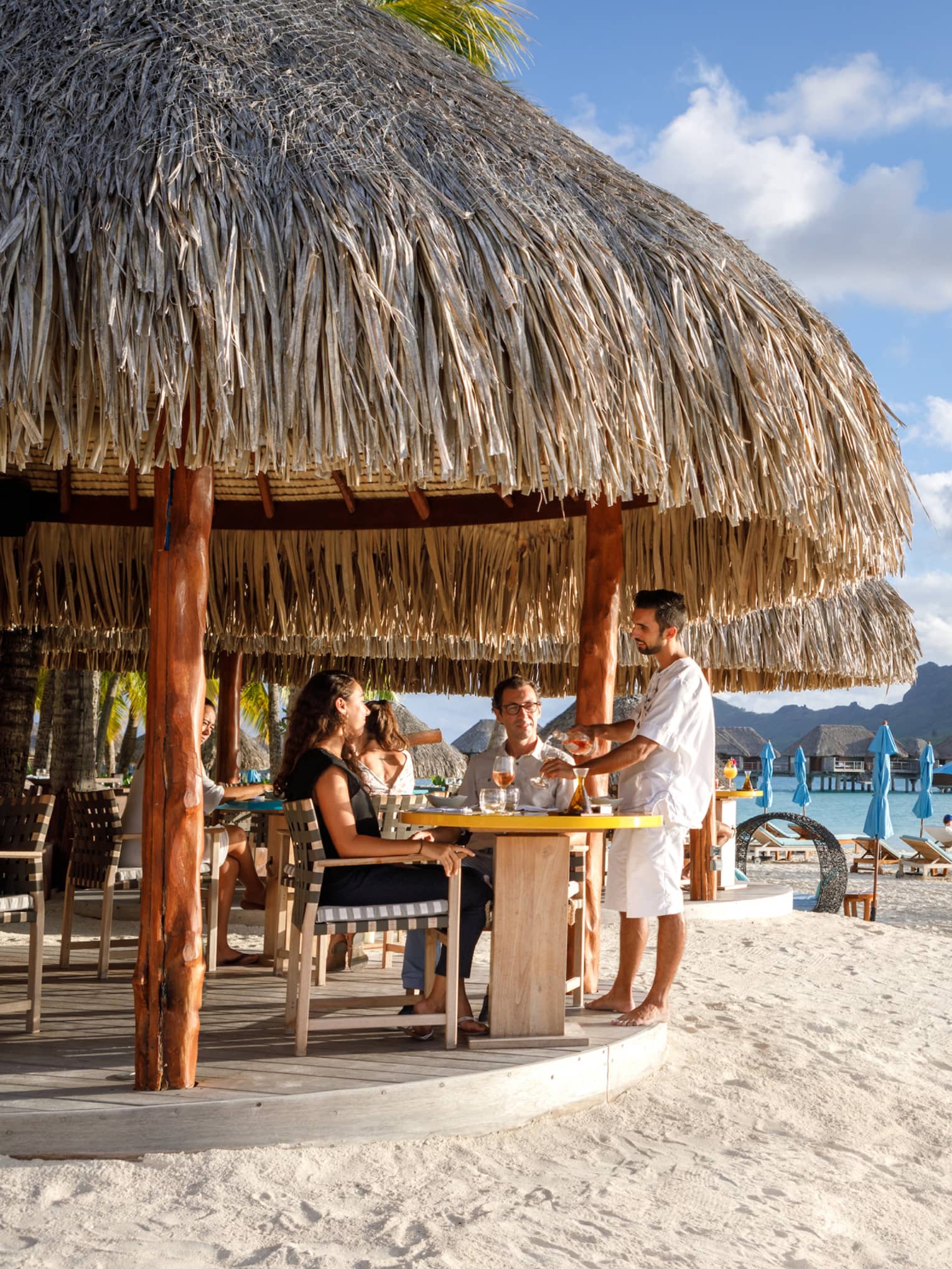 Couple seated in bar under grass roof on beach, view of people in personal cabanas, mountain view