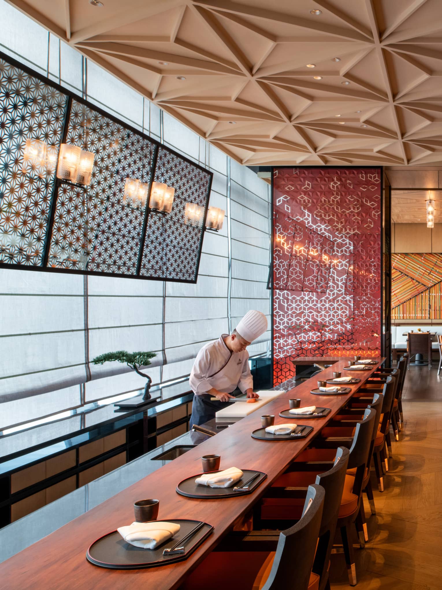 A sushi chef works by a long wood dining table in a modern restaurant, a red ornate screen and walls reflecting golden light.