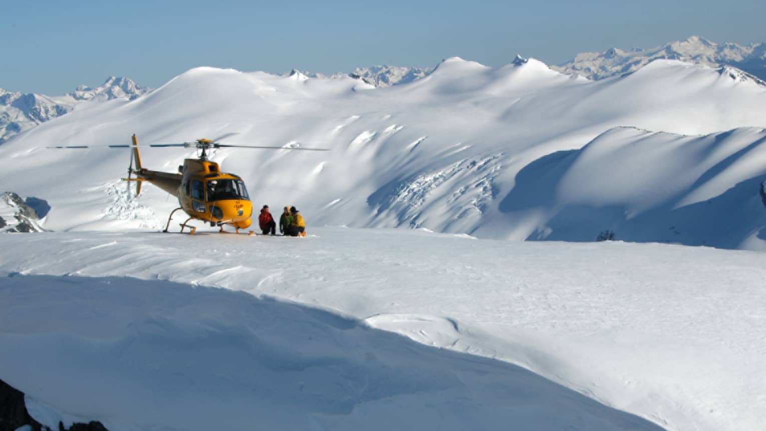 Group in snowsuits stand beside yellow helicopter landed on snowy glacier 