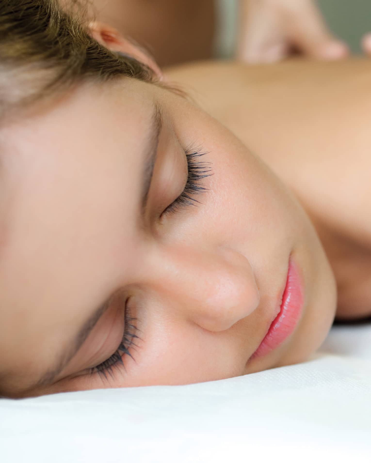 Woman with eyes closed rests on spa treatment table while massage therapist places hands on her bare back