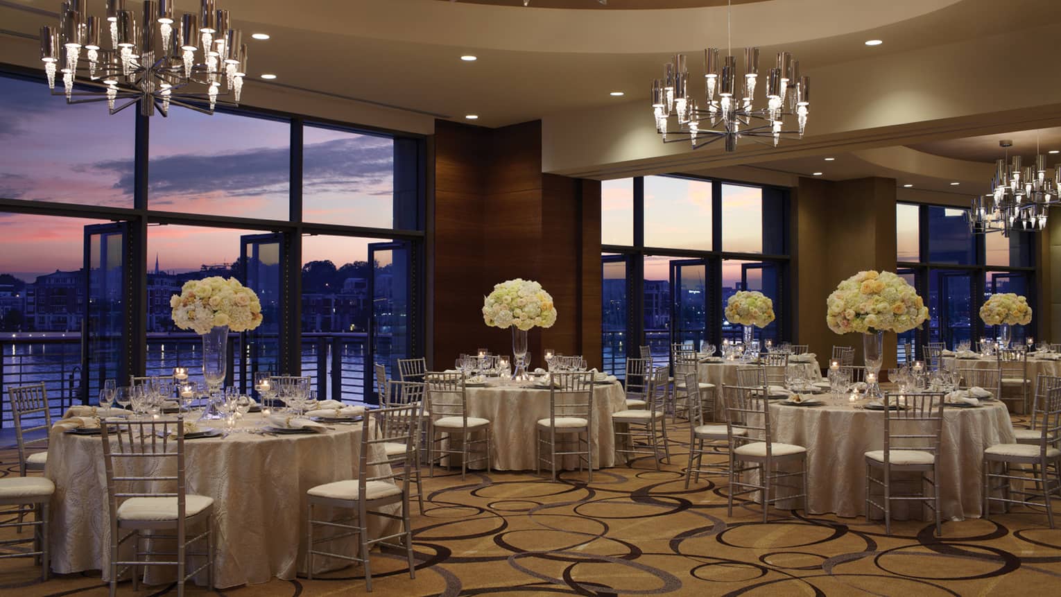 Ballroom banquet dining tables under chandeliers and large windows with sunset Harbor views 