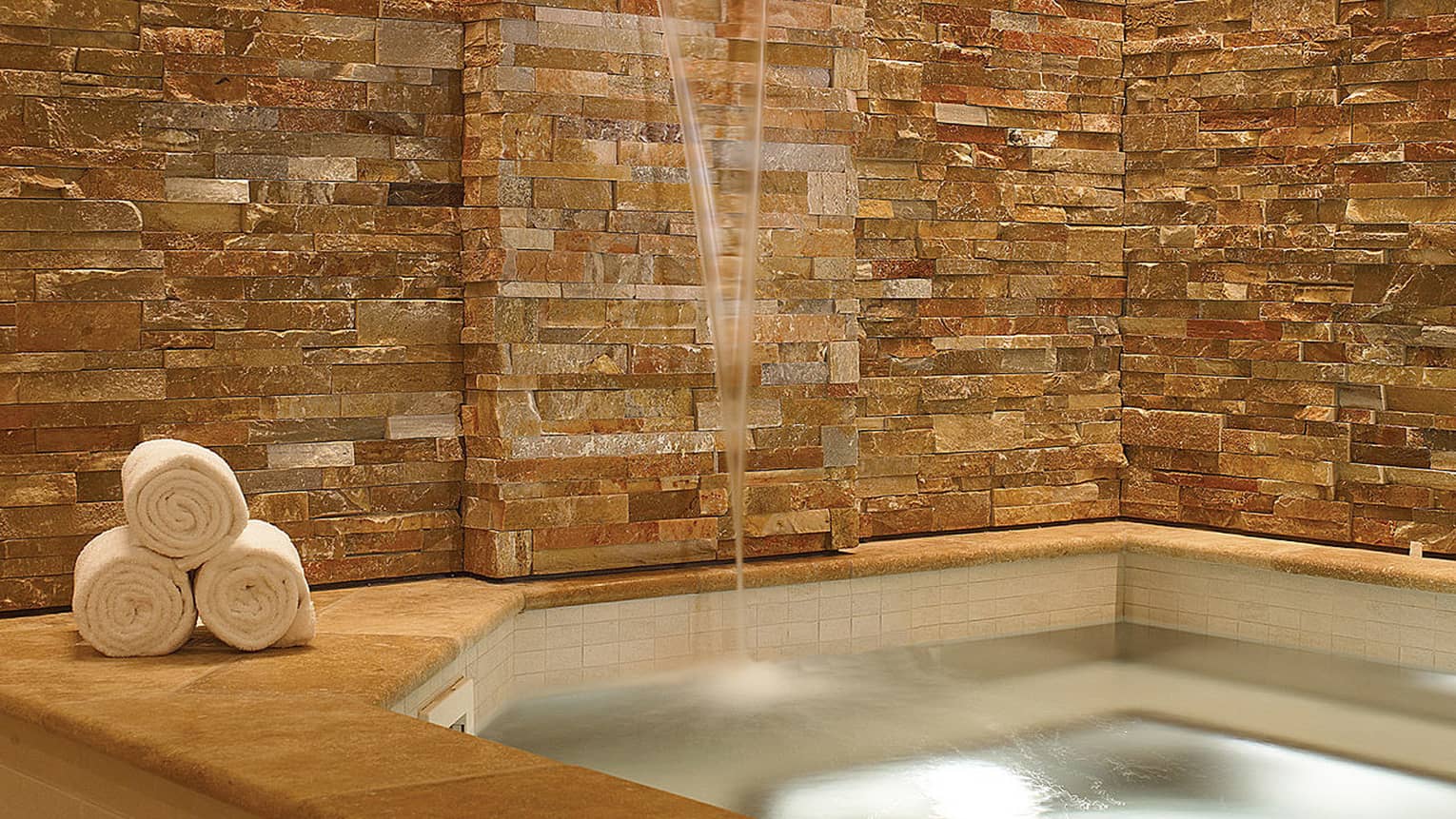 Glowing spa tub surrounded by stone wall and three rolled white towels on ledge