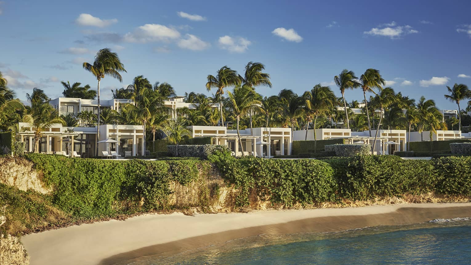 Modern-looking white ocean villas with tall palm trees perched on grass, white sand beach