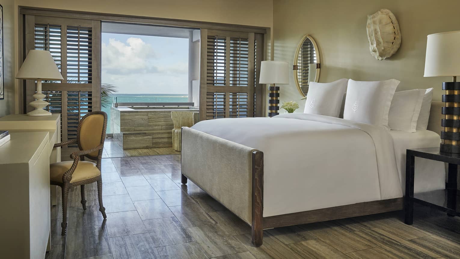 Elegant bedroom with large bed, desk and chair, wooden floors and ocean view through wide windows with shutters
