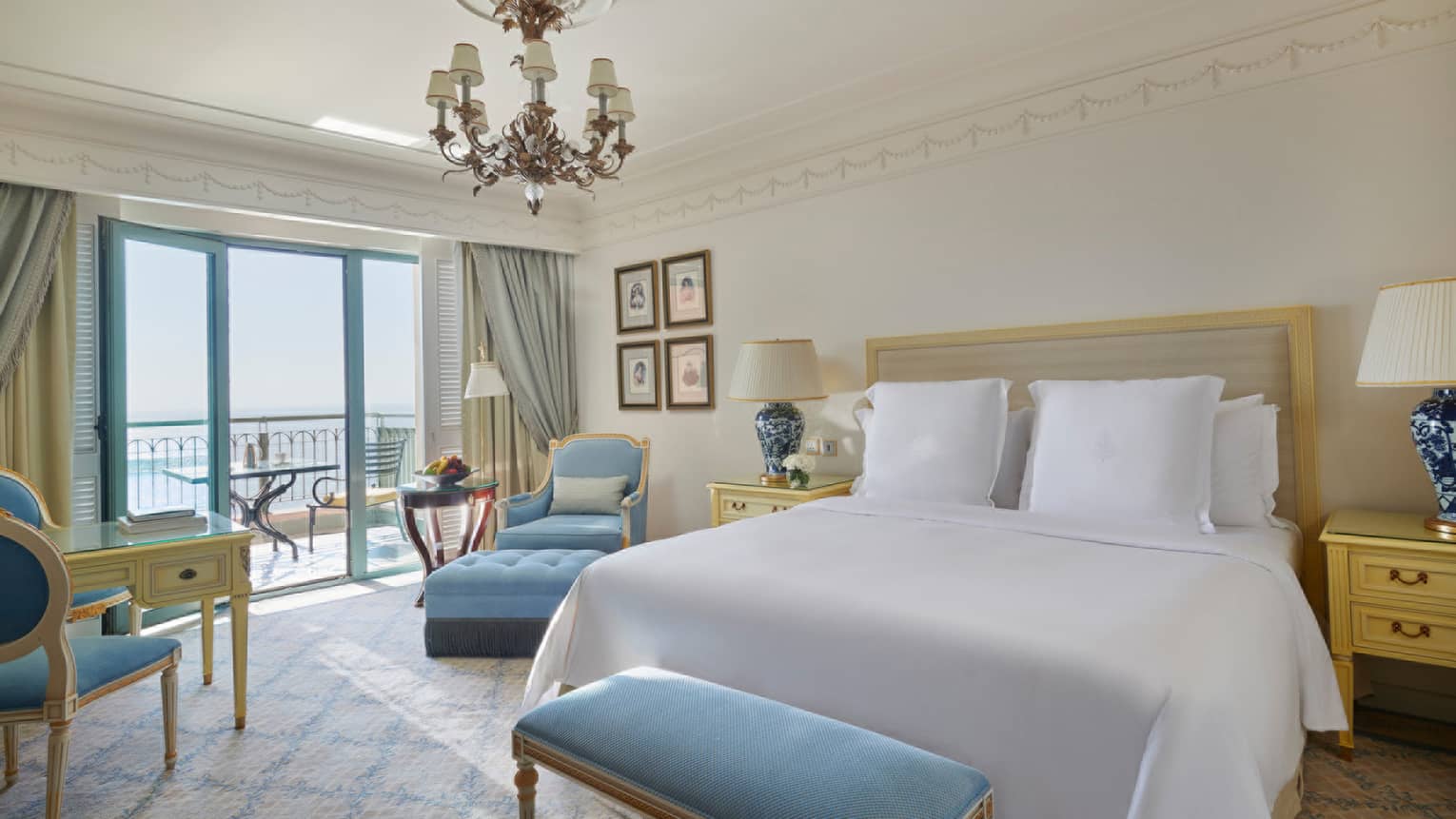 Bedroom with white king bed, chandelier, light blue bench and arm chair, and a balcony overlooking the sea