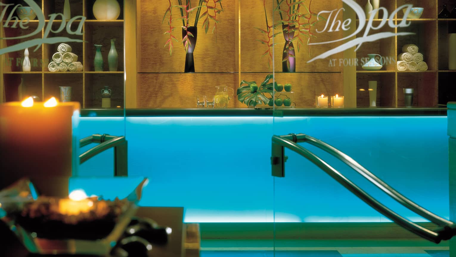 Glass doors at spa lobby, reception desk with with glowing candles, rolled up white towels on shelf 