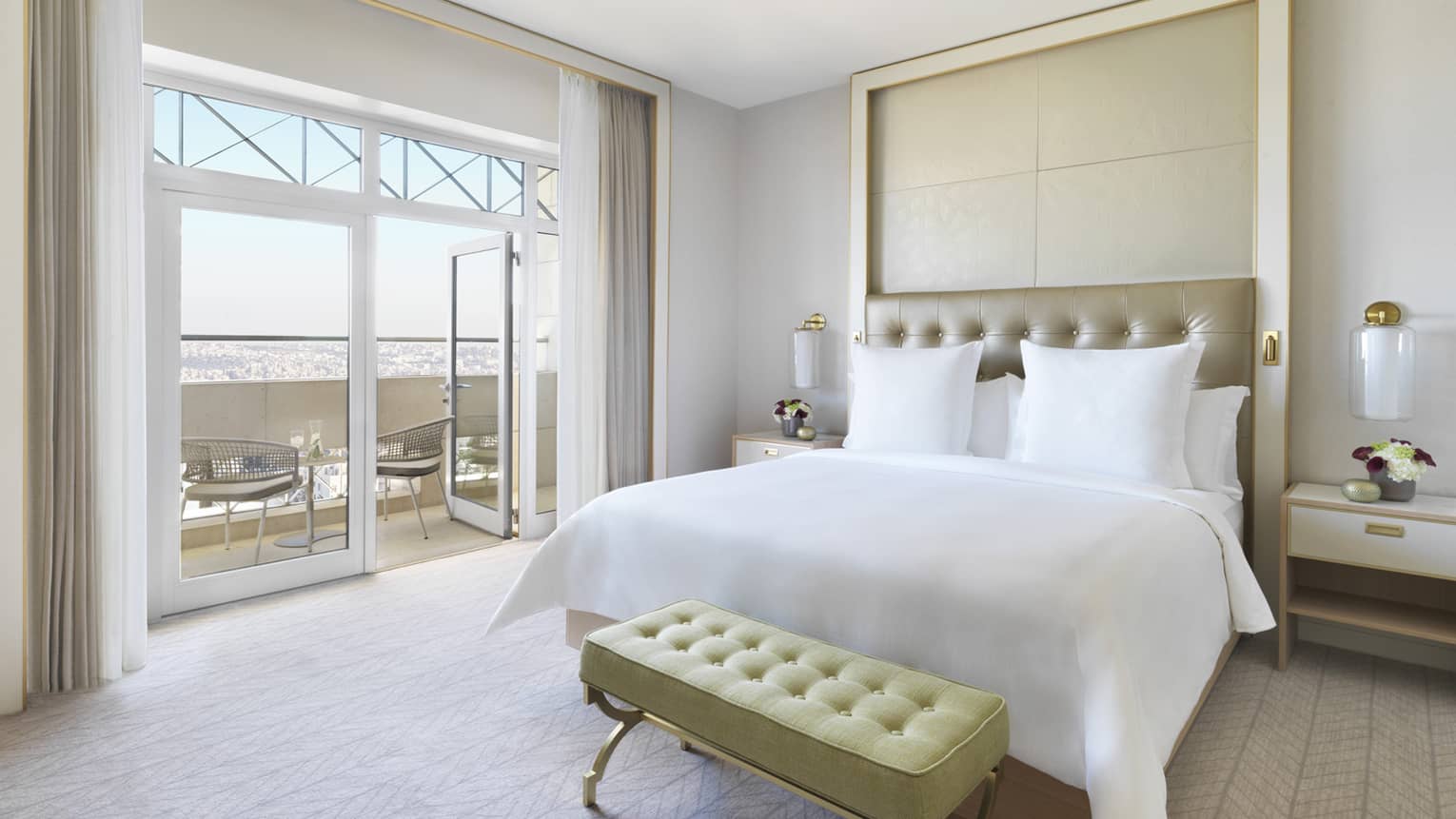 A plush white bed in the centre of the room with a yellow ottoman at the foot. The room opens up to a veranda overlooking the city. 