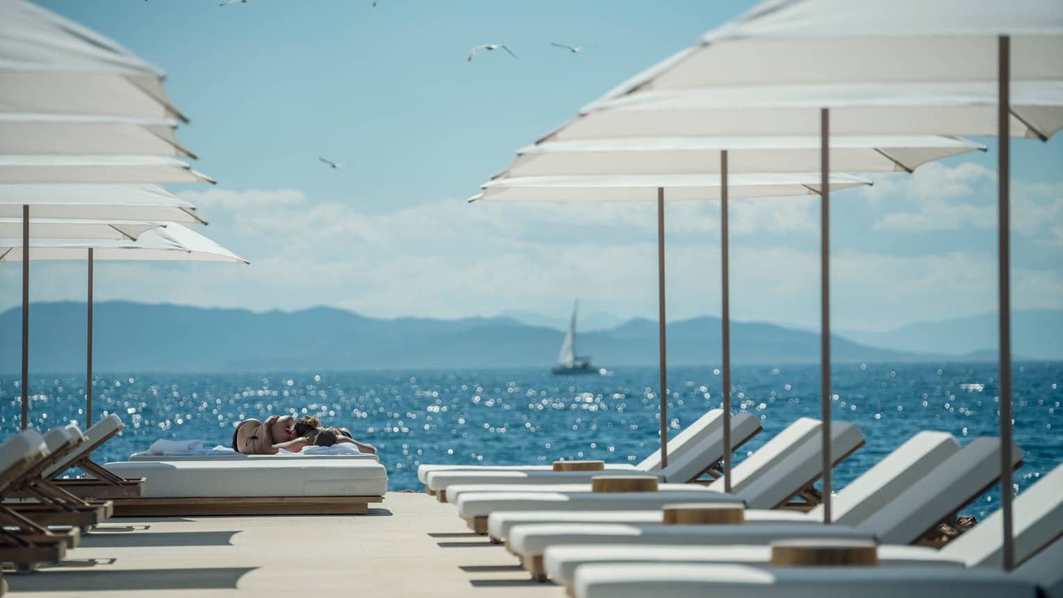 A sail boat sails in the distance while white lounge chairs are ready for guests and lined up next to the oceanside Nafsika Pool in Athens. 