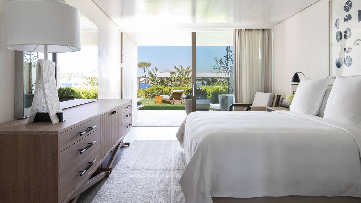 Bedroom with plush king bed, modern wooden dresser with lamp, walk-out terrace to garden