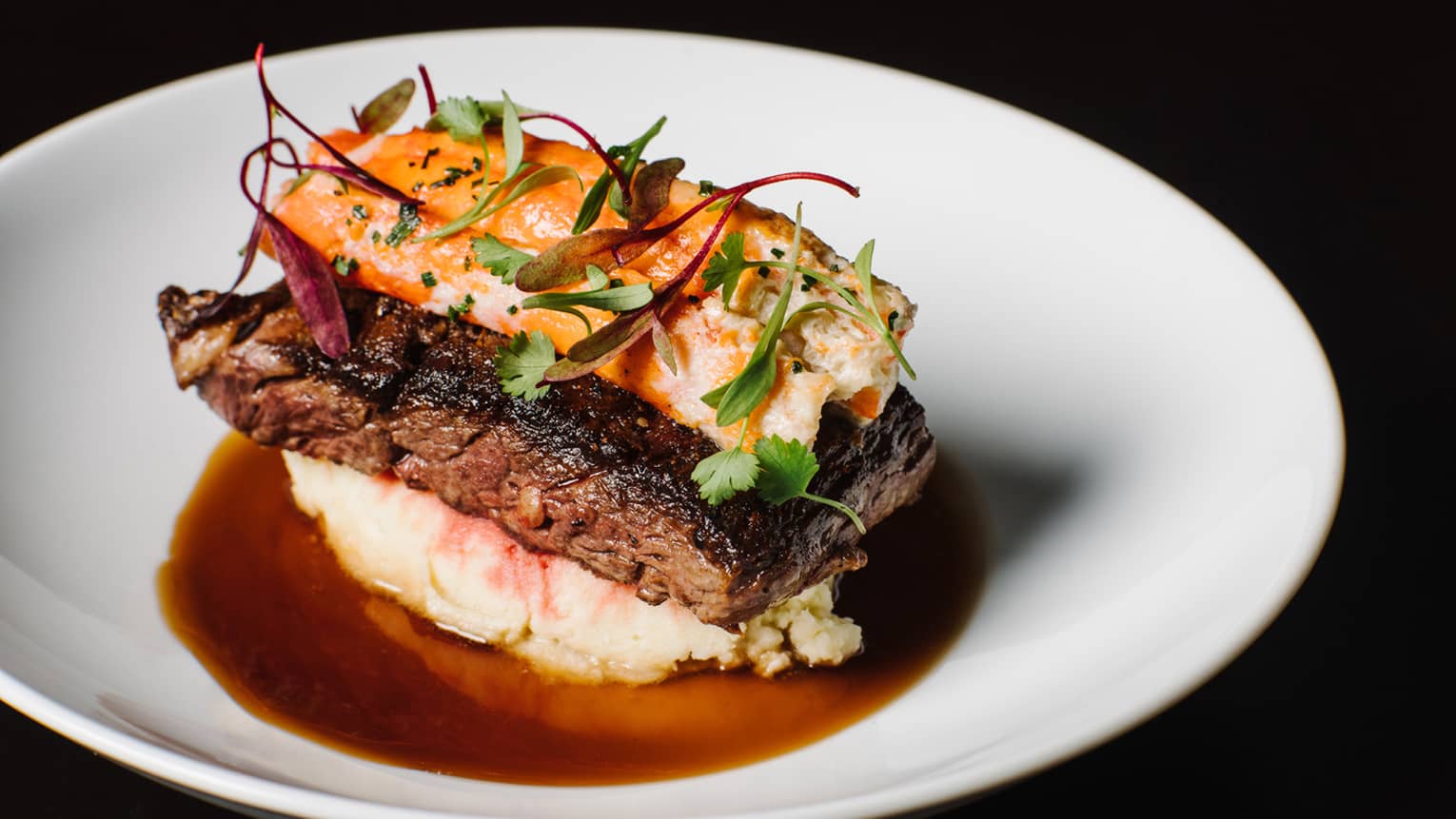 Close-up of grilled steak topped with lobster and fresh herbs, on bed of mashed potato with jus