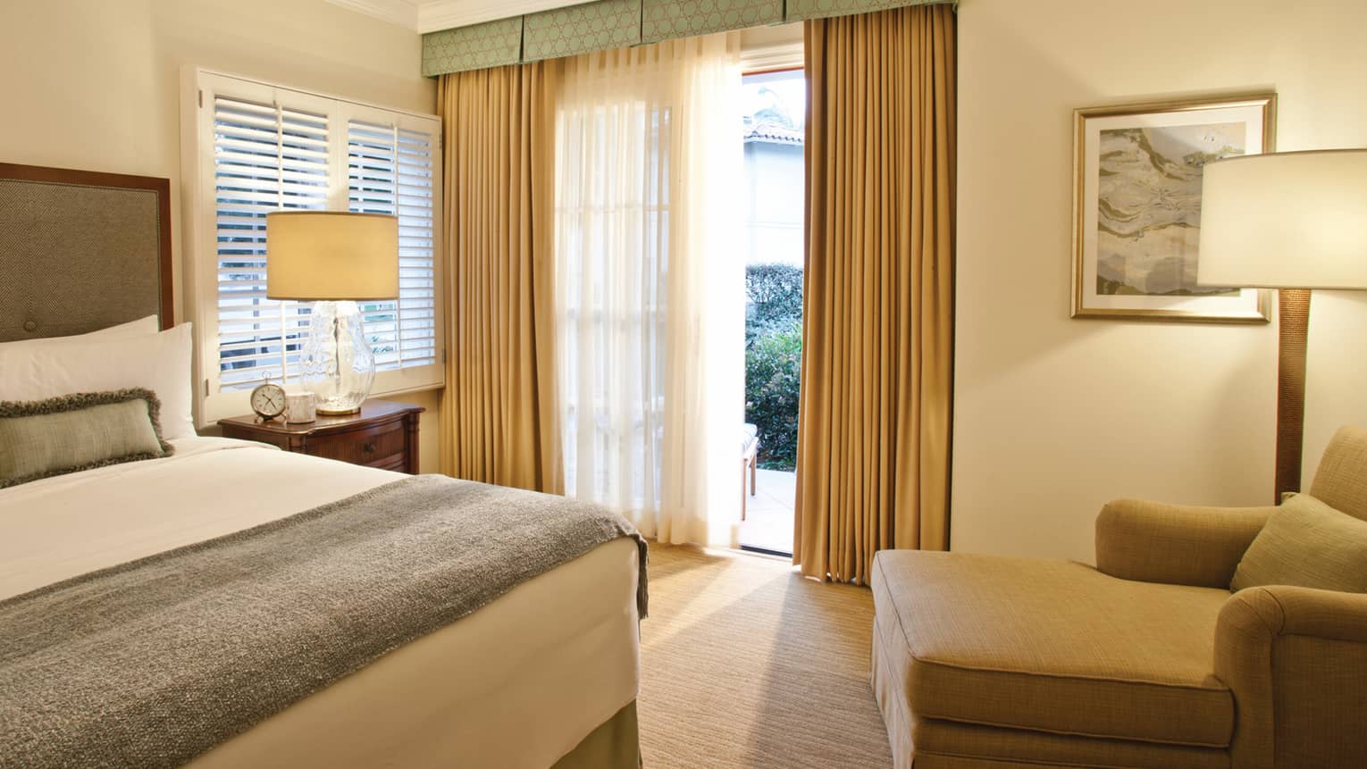 Residence master bedroom with corner of bed, green blanket and pillows, open patio door with long curtains, armchair
