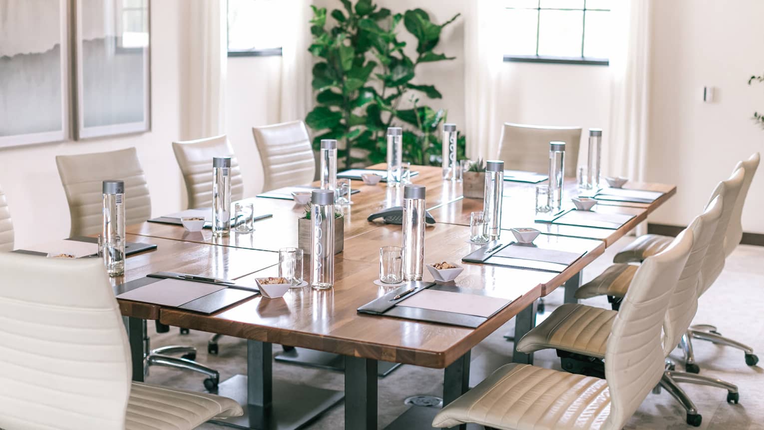 A rectangular boardroom table set with clear glass water bottles and black folders in a naturally-lit room