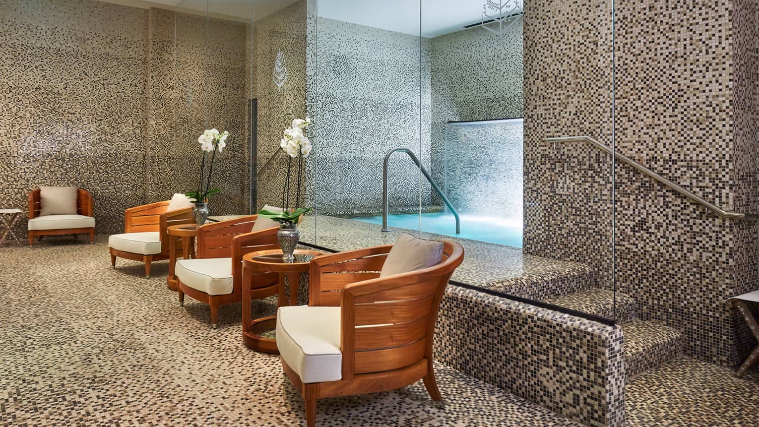 Tile spa room with whirlpool bath behind row of wood chairs with white cushions, tables with white orchids