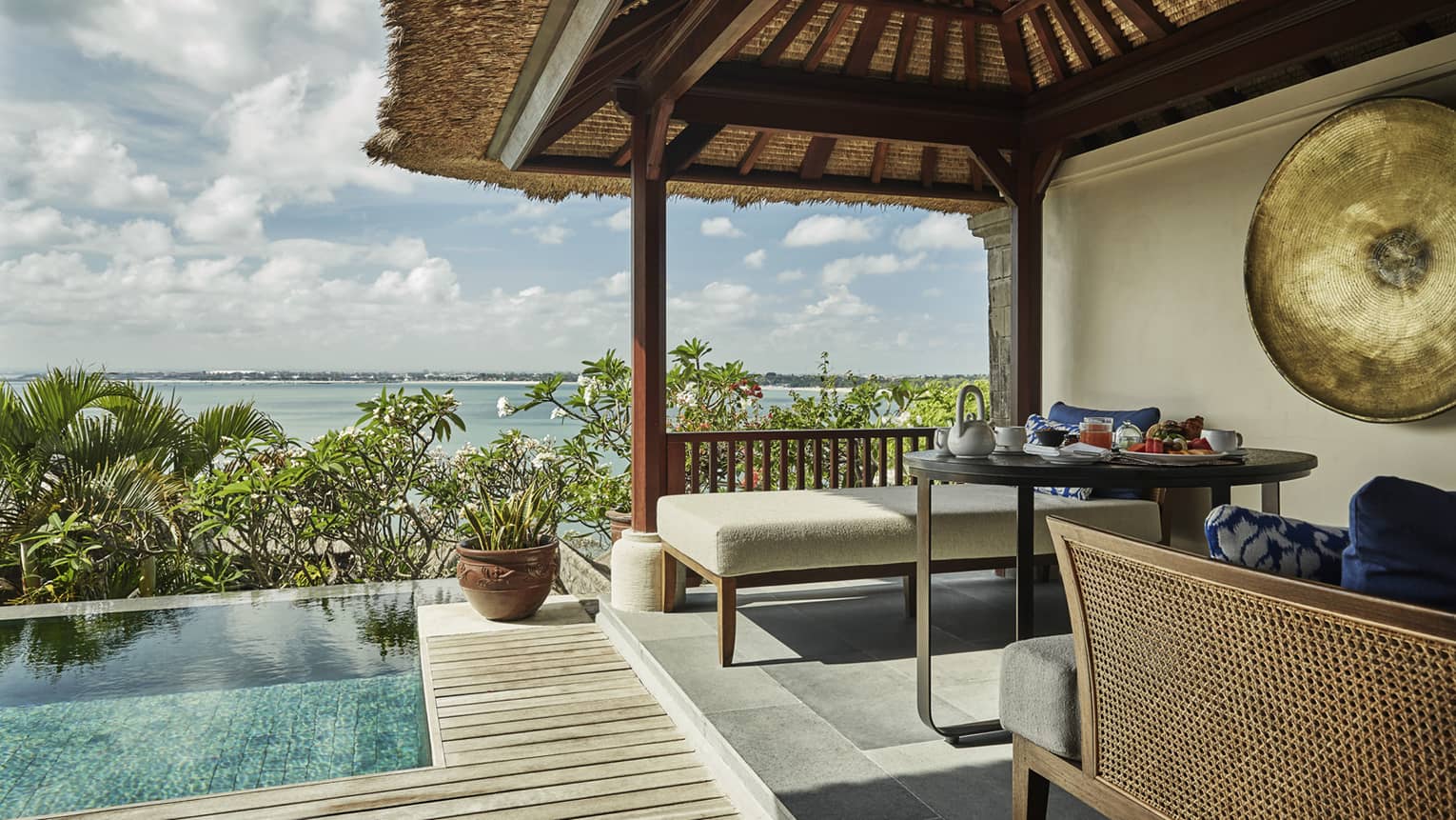 Close-up of Deluxe Villa patio corner with swimming pool, plant, white bench under thatched roof, ocean view