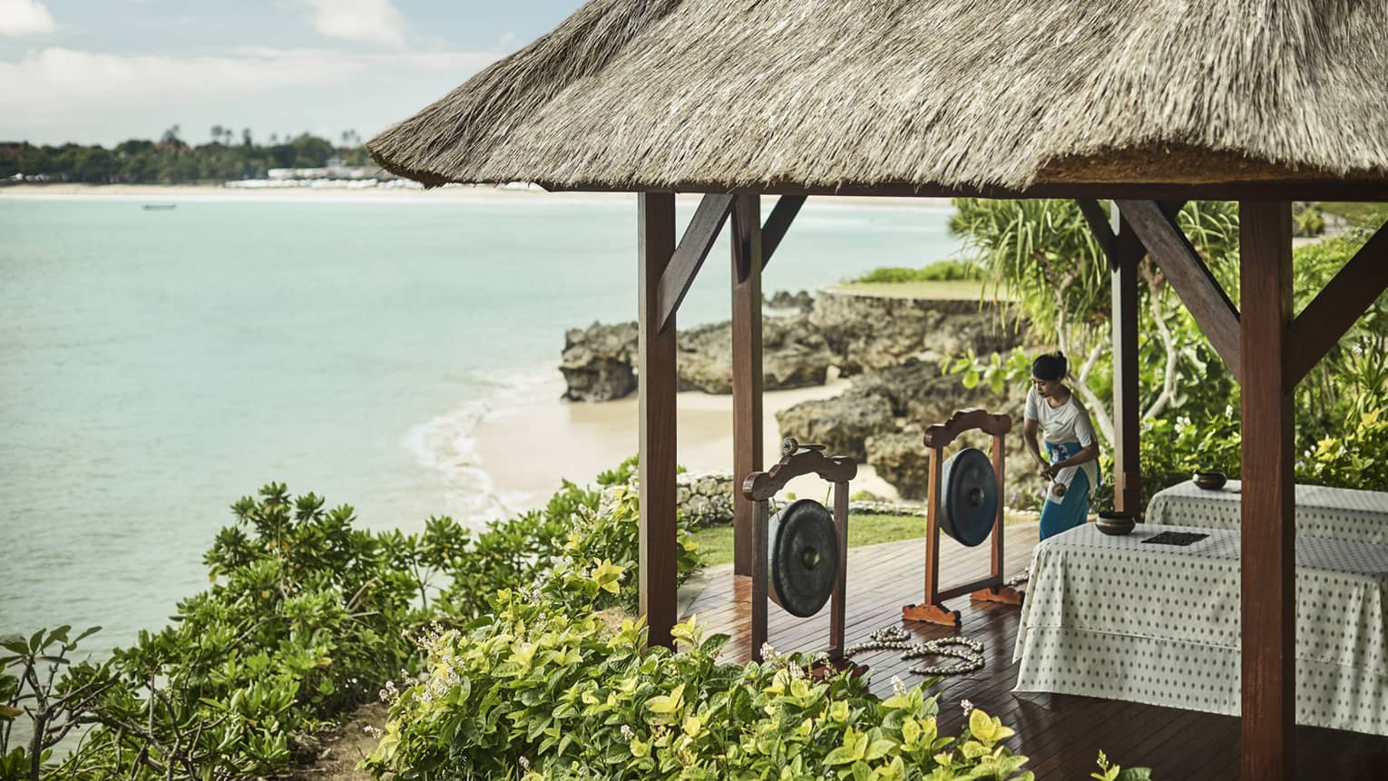 Spa attendant prepares to strike Balinese in front of two massage tables under gazebo overlooking beach, ocean