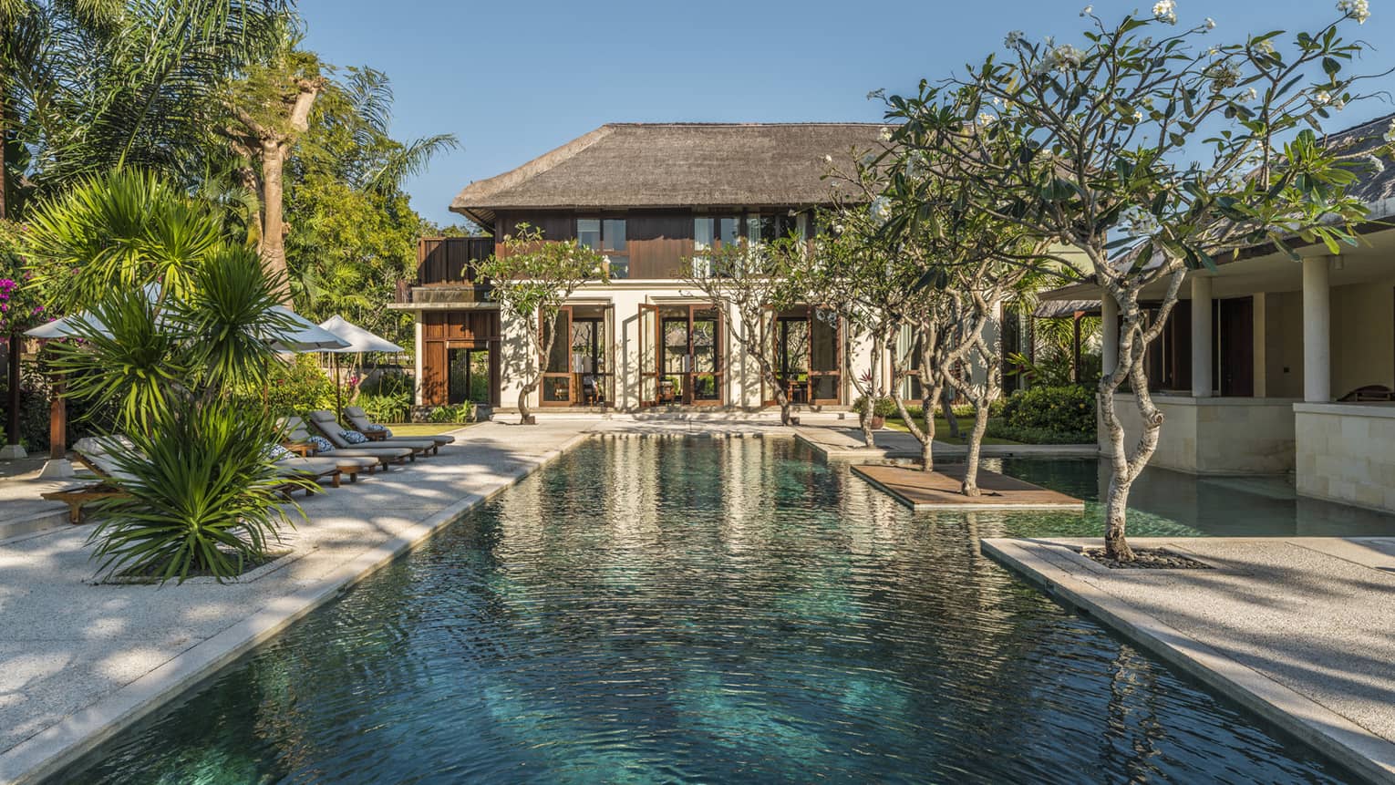 Two-storey villa at end of long outdoor swimming pool lined with trees, tropical plants