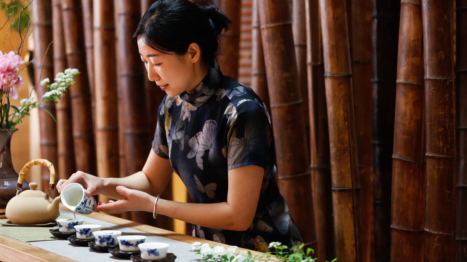 Woman pours tea into small cups in front of bamboo wall