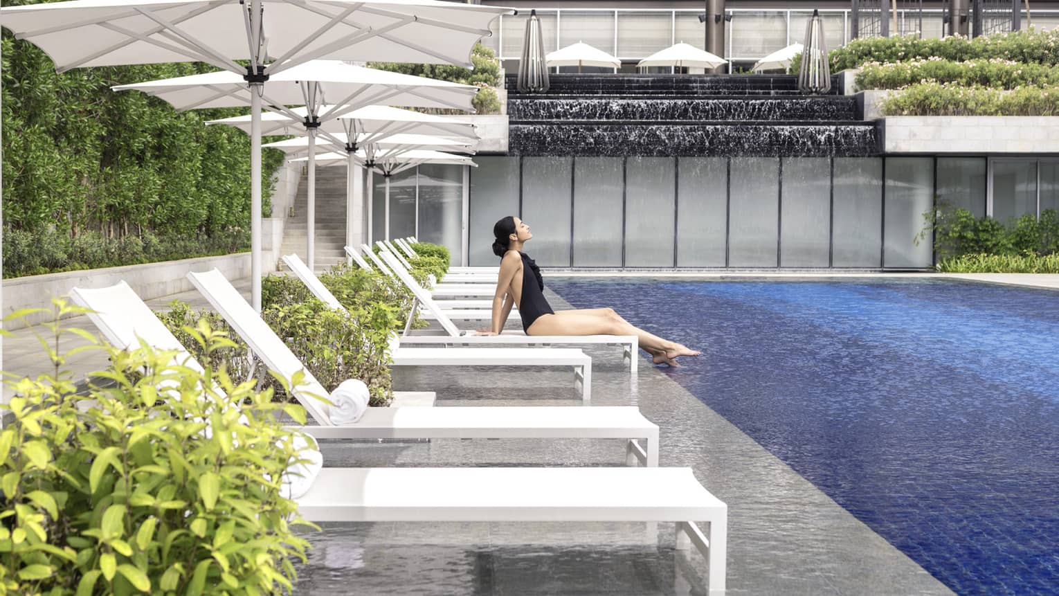 A woman in a black bathing suit relaxes on a chaise by the pool