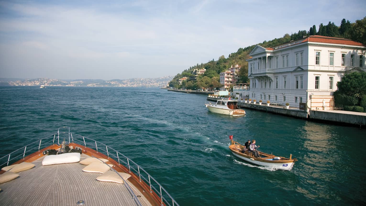 View from deck of yacht, small boat passes by on sea, waterfront path, green hill