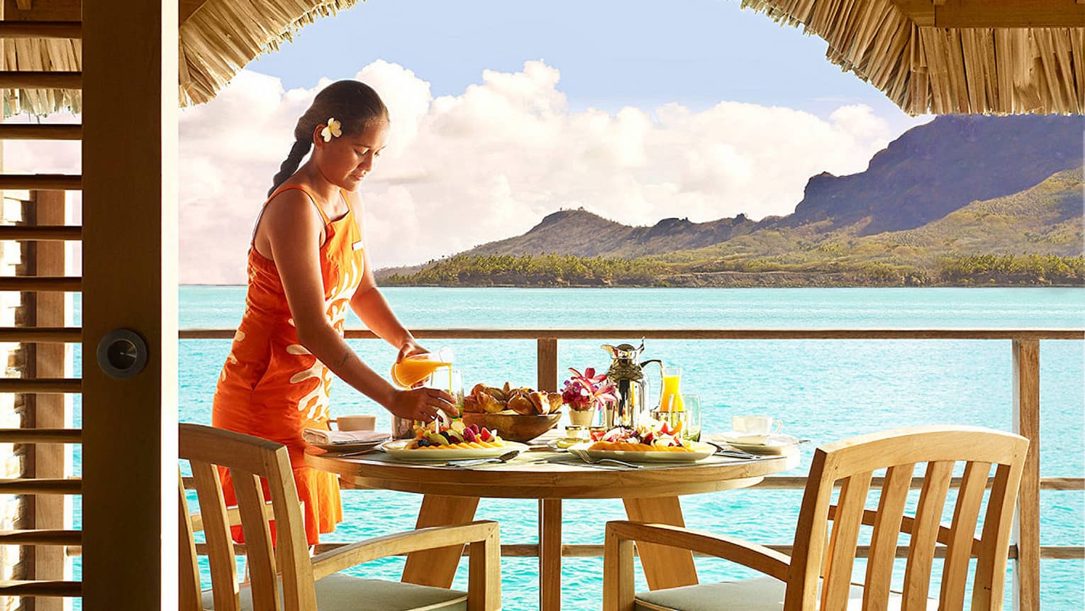 Woman in orange dress and gardenia behind ear pours orange juice at breakfast dining table on patio