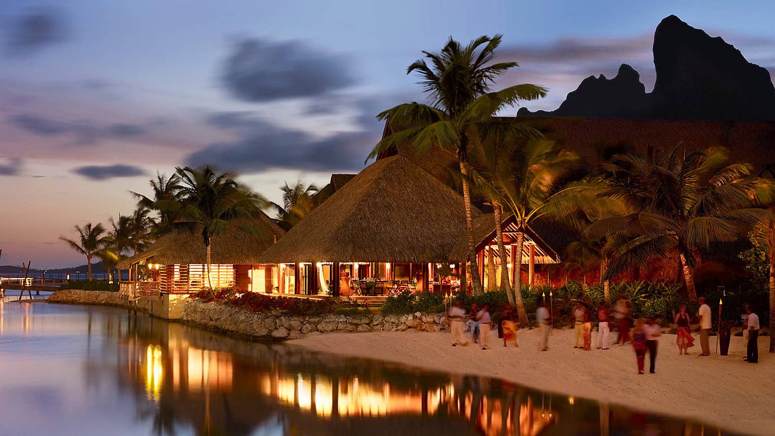 Guests on sand beach by pavilions with lights at night